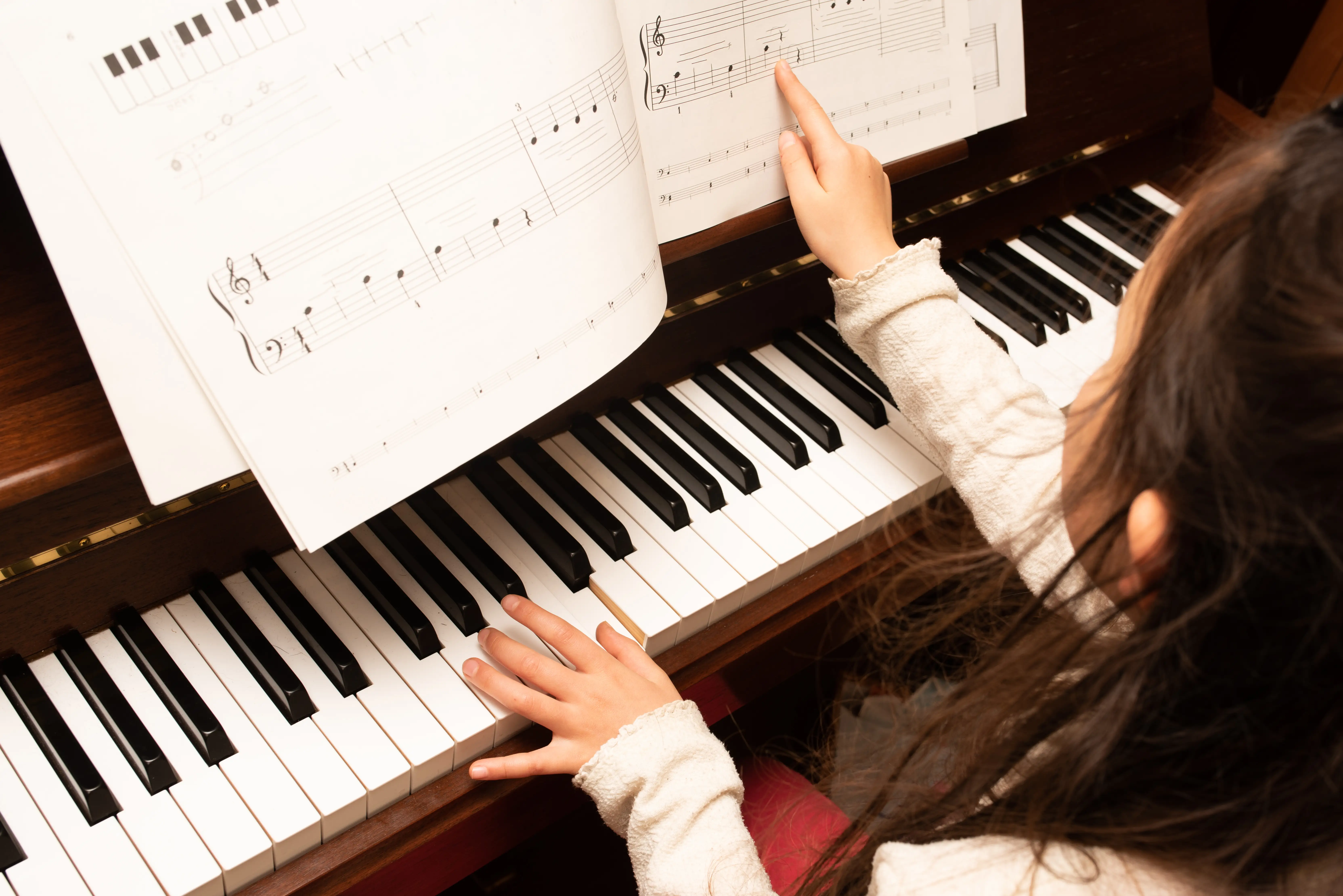 Person at an upright piano points to sheet music while one hand rests on the keyboard.