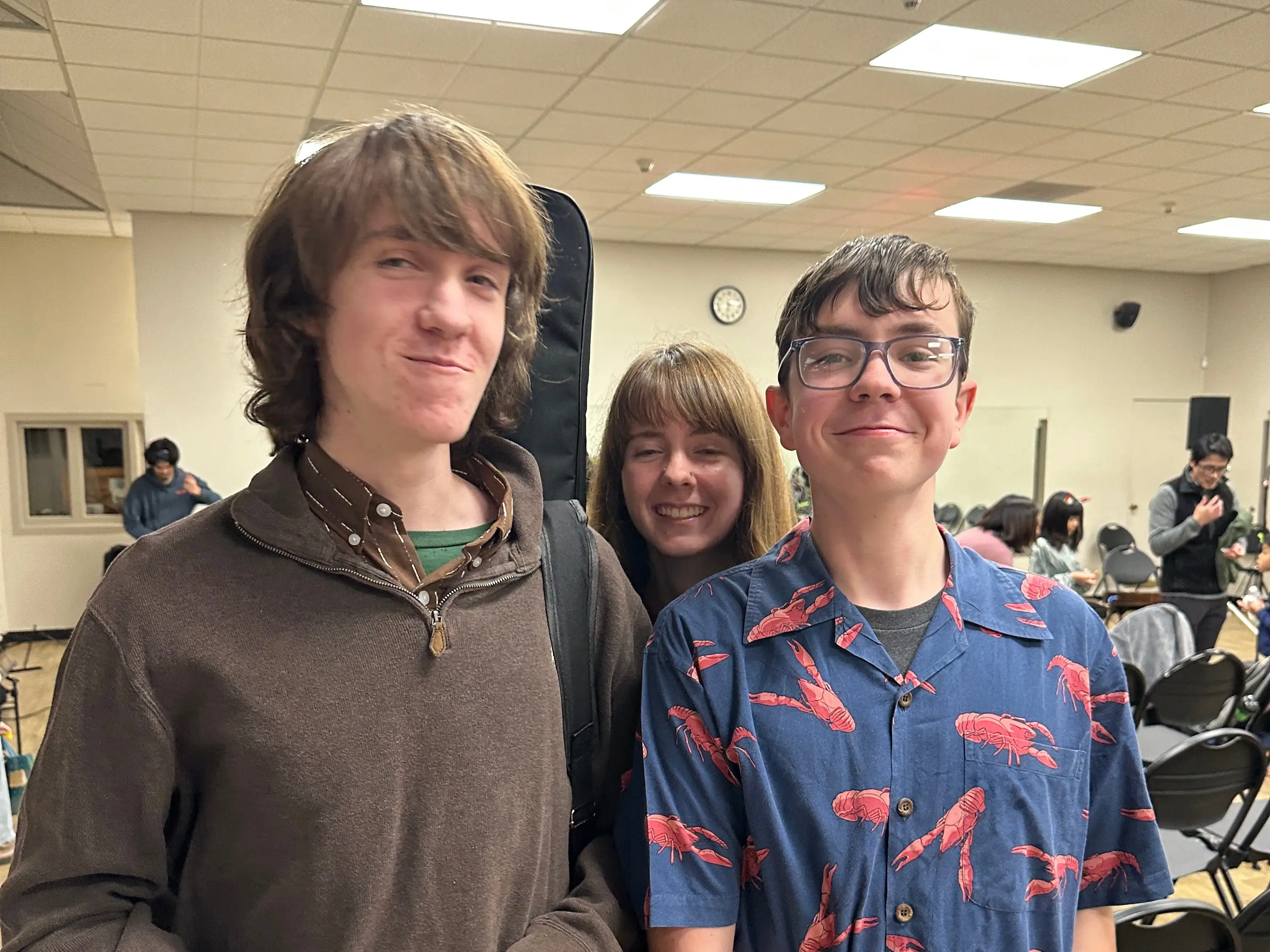 Three smiling people stand together in an indoor room with folding chairs and fluorescent ceiling lights.