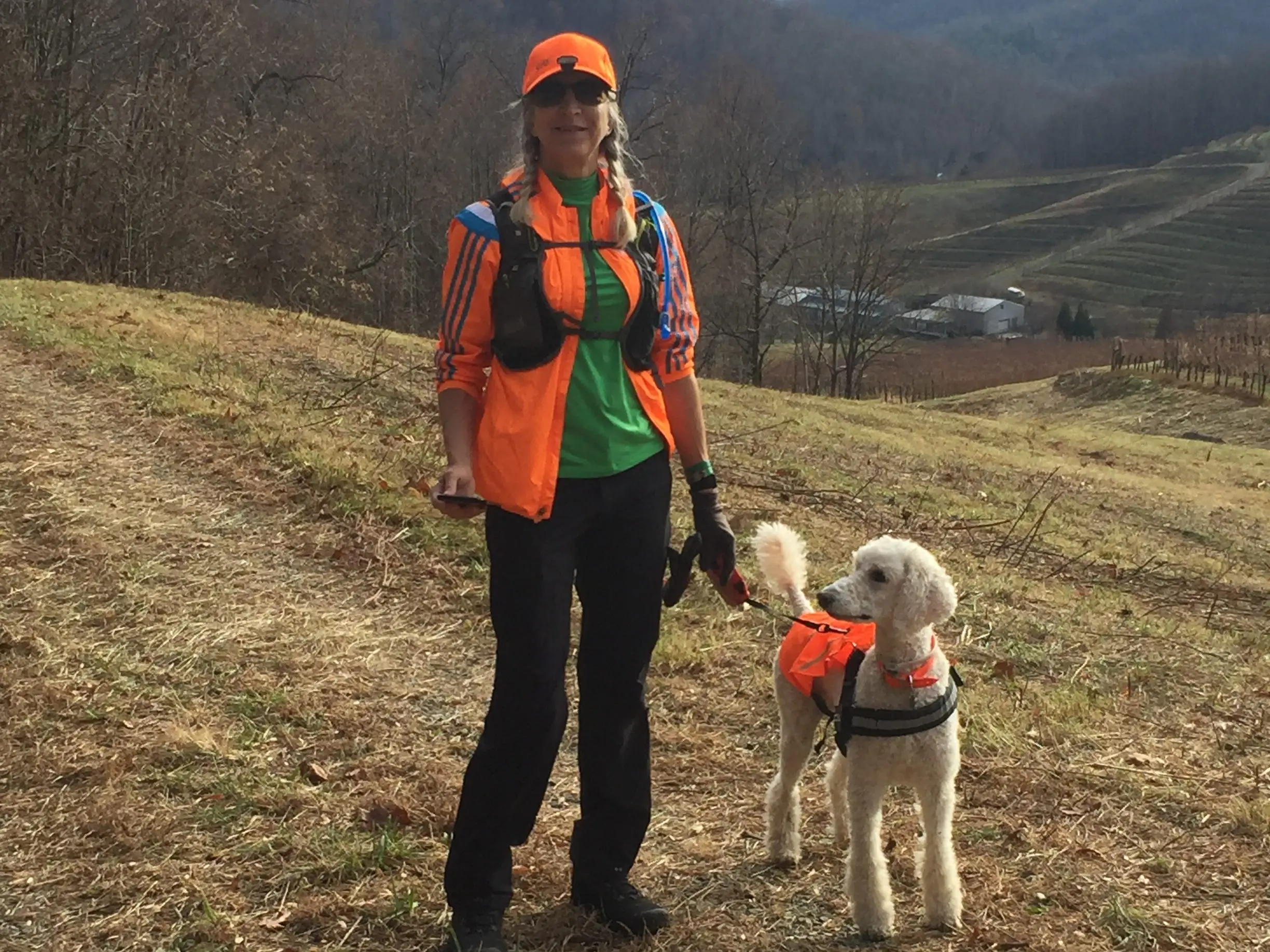 Person in bright orange outdoor gear stands with a white dog on a hillside overlooking wooded mountains.