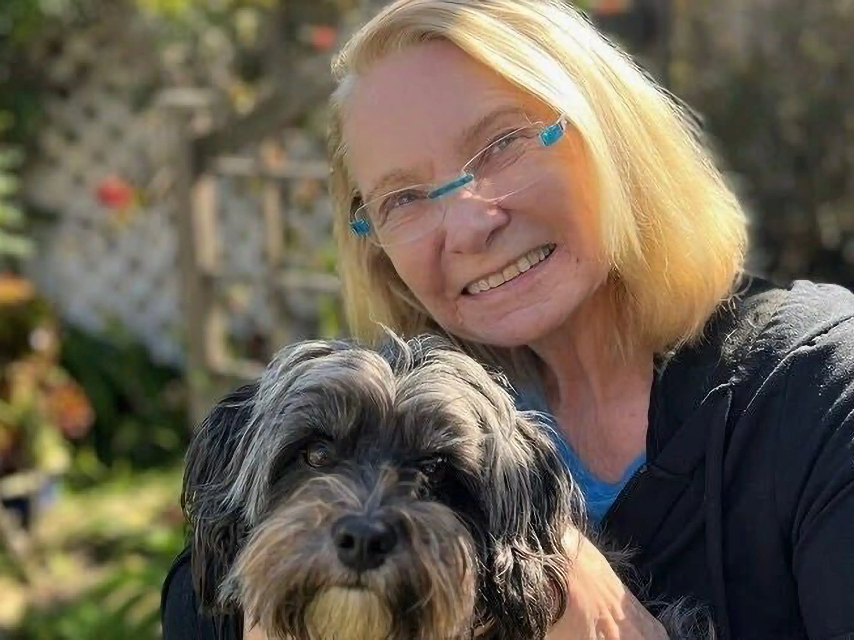 Smiling person with glasses holds a shaggy black and gray dog in a sunlit garden.
