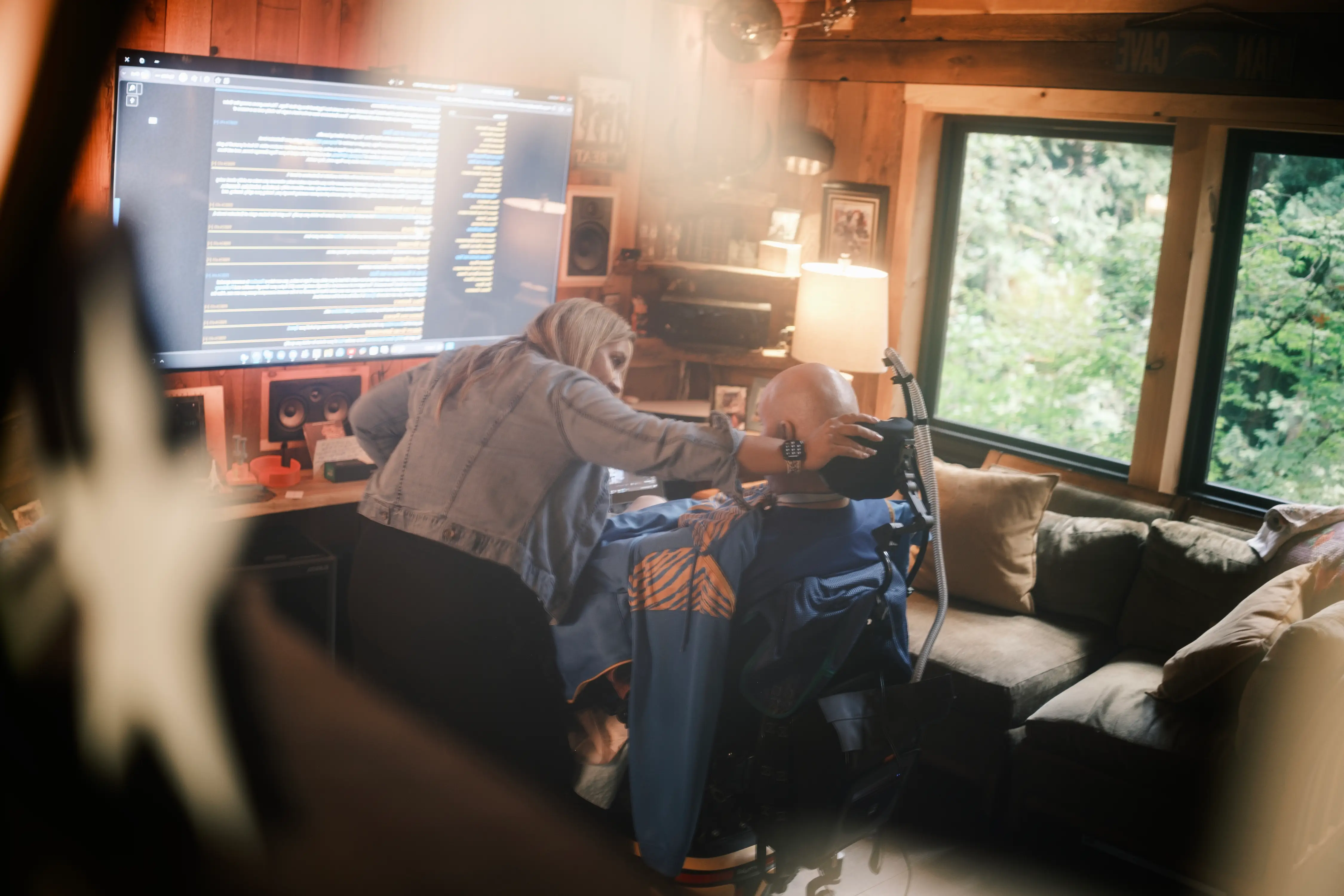 Person leans over a seated individual using head-mounted equipment beside a large monitor in a wood-paneled room.