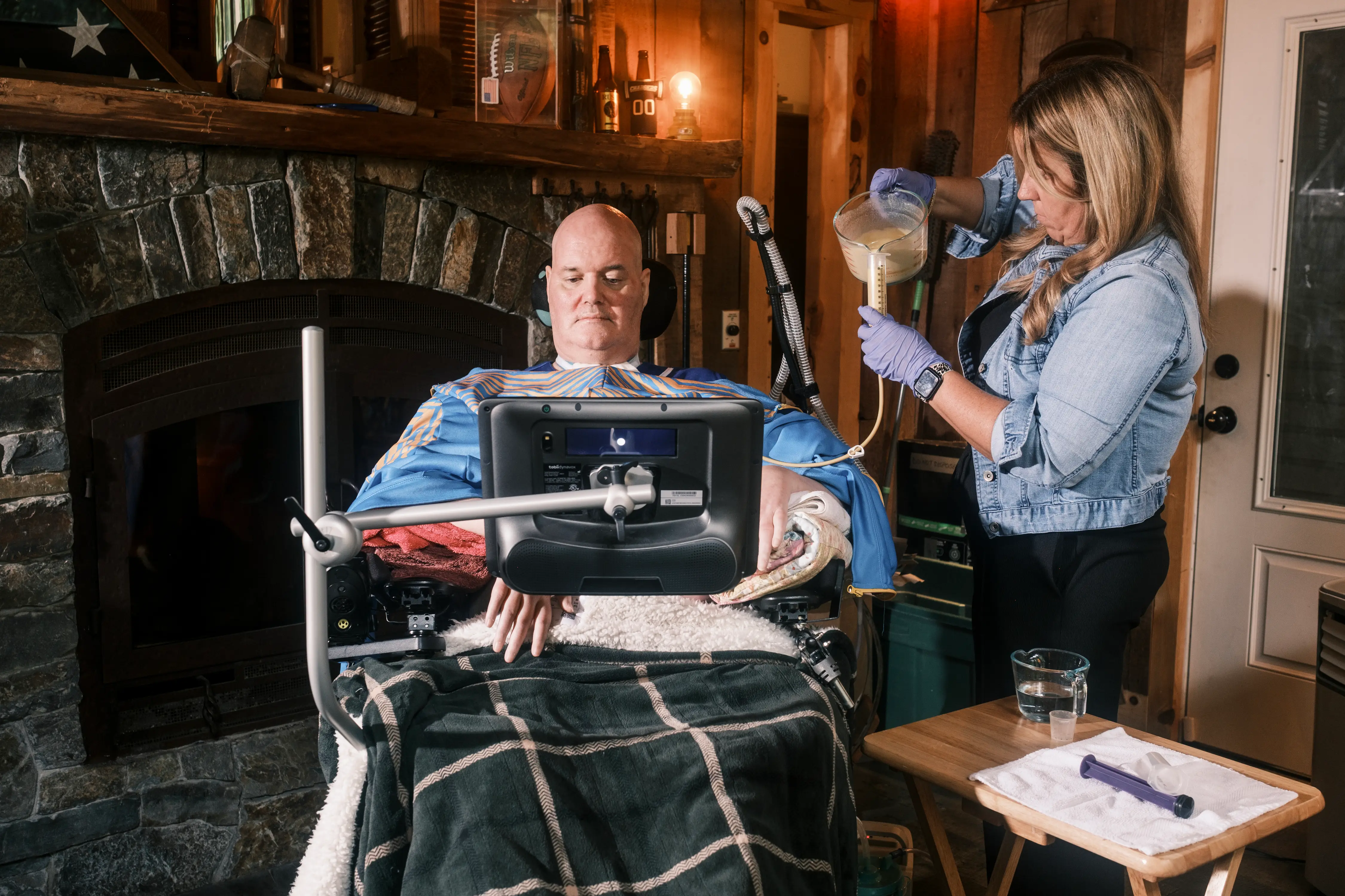 Caregiver in gloves pours liquid nutrition into a tube feeding bag beside a seated person using a mounted device.