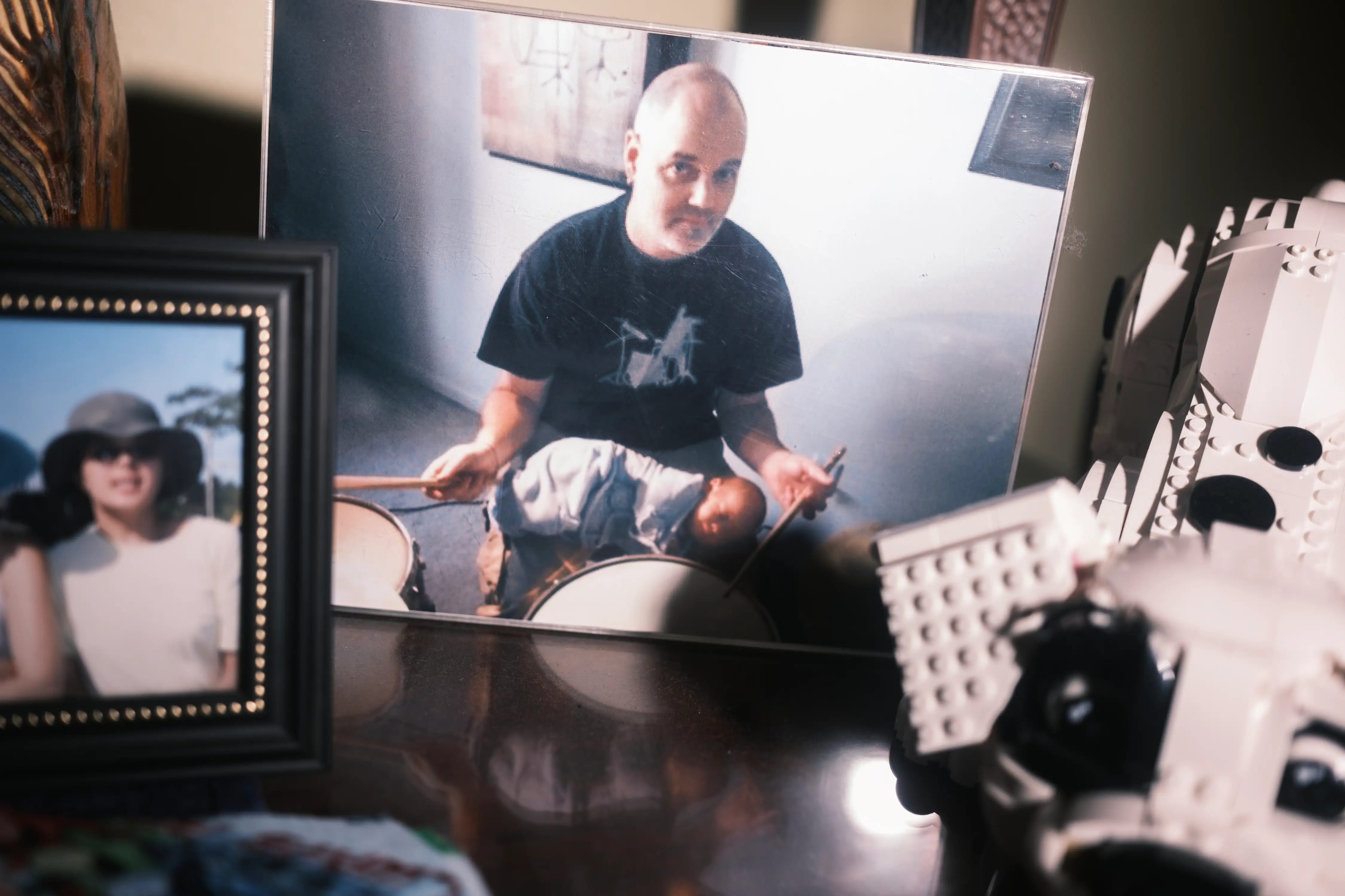 Framed snapshots, including a person playing drums with a child, sit beside a white toy brick model.