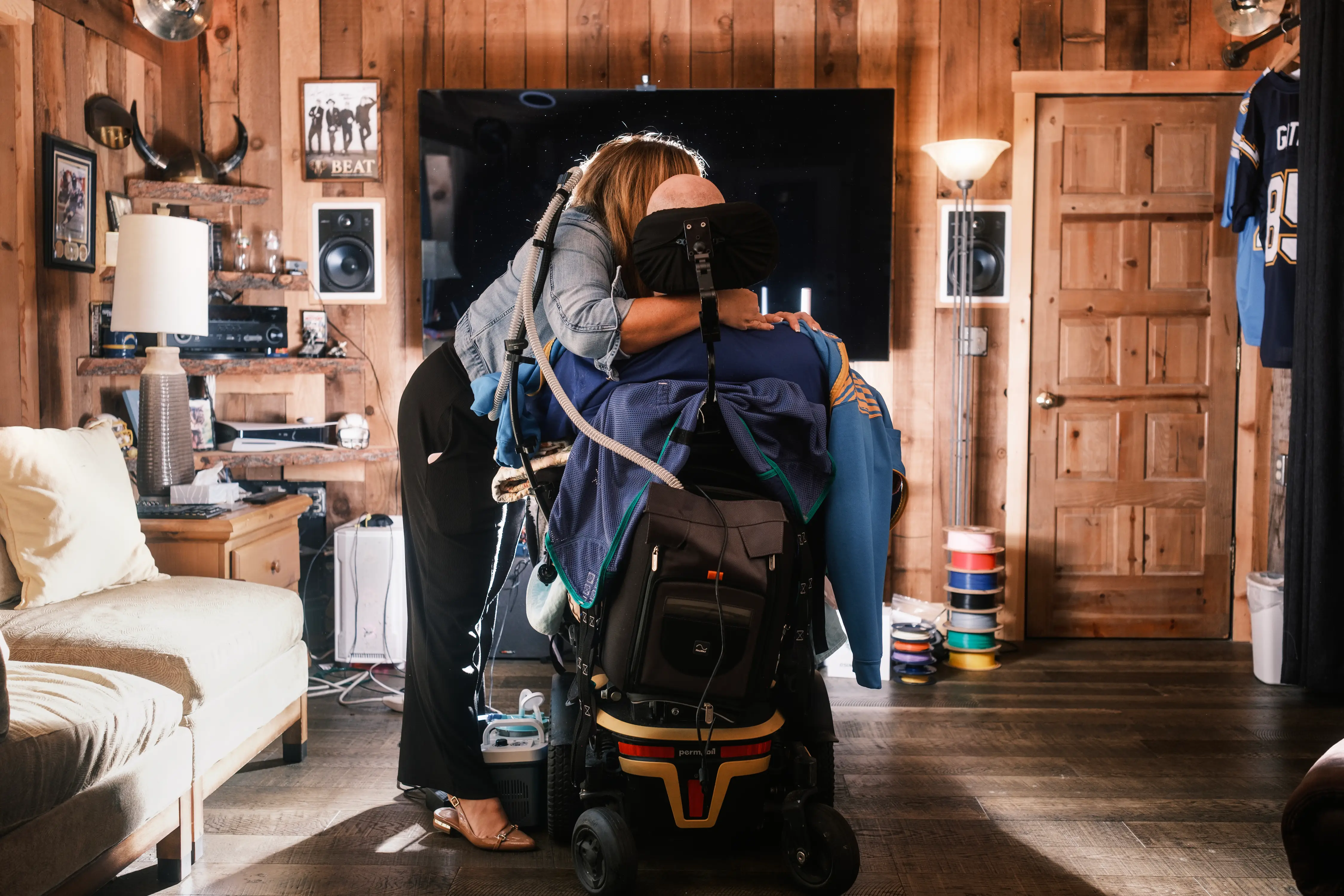 Two people embrace in a wood-paneled living room, one seated in a powered wheelchair with attached tubing.