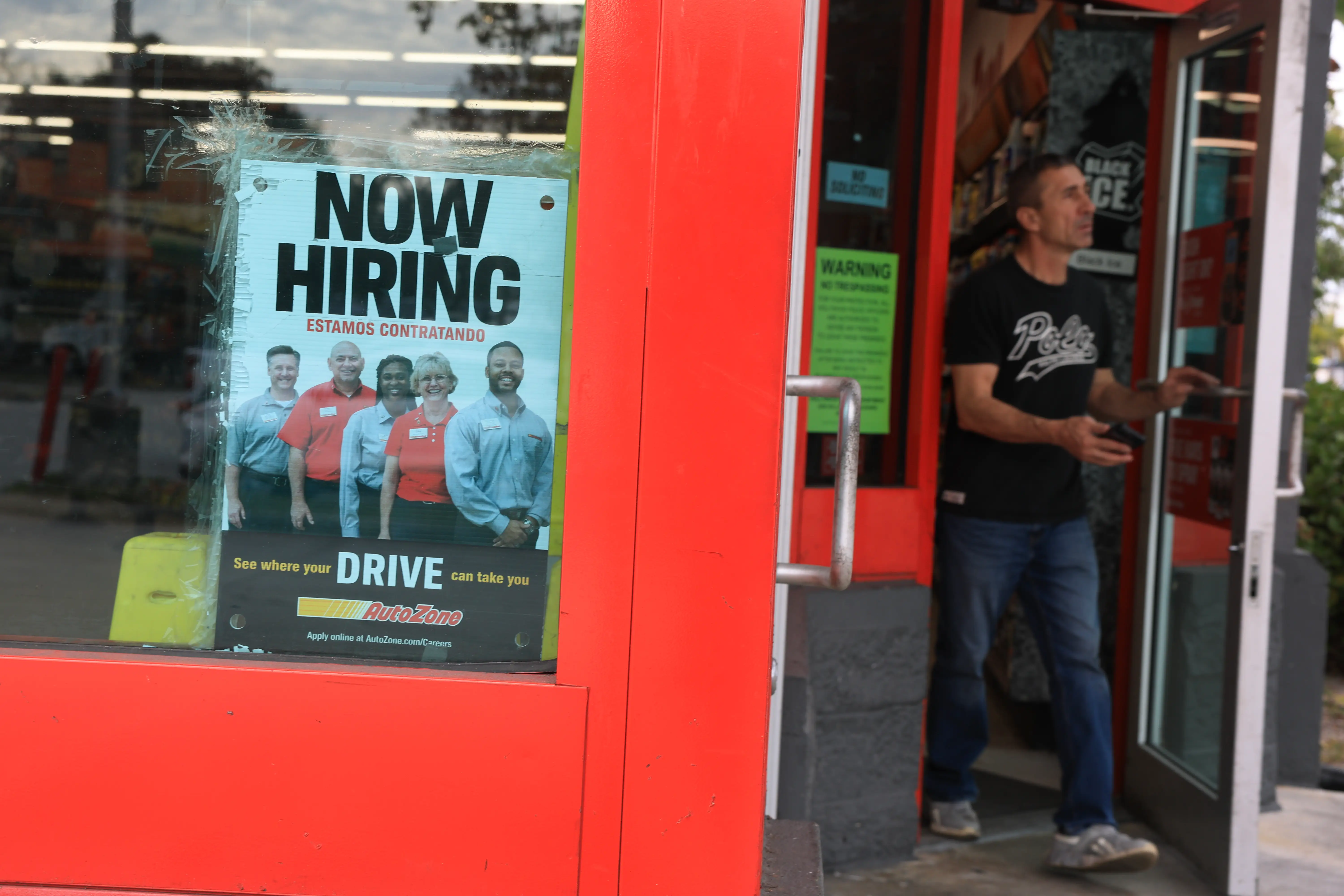 AutoZone storefront entrance with a “NOW HIRING” sign in the window and a person walking out.