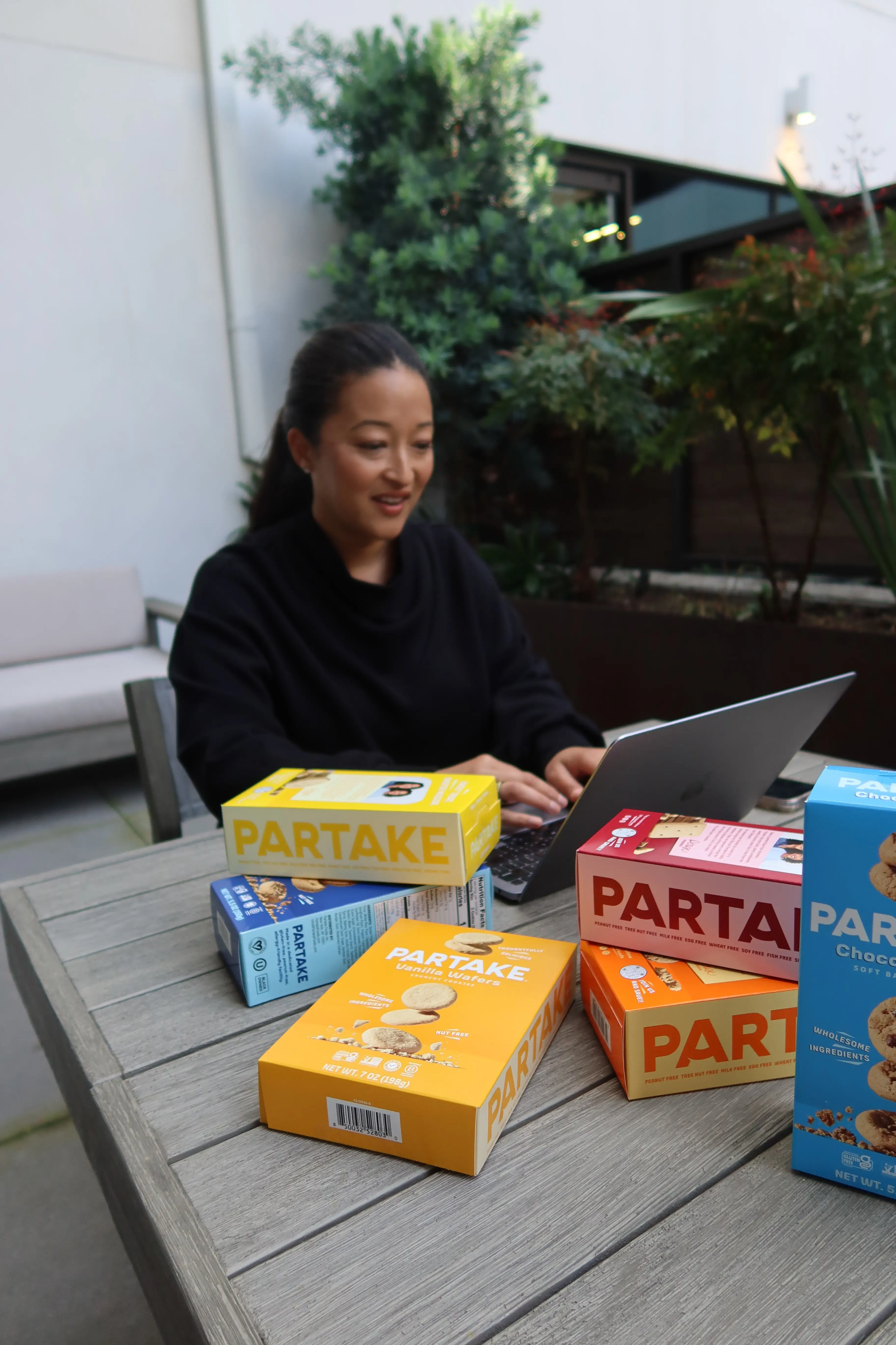 Denise Woodard working on her laptop at an outdoor table with Partake cookies packaging scattered on the table.