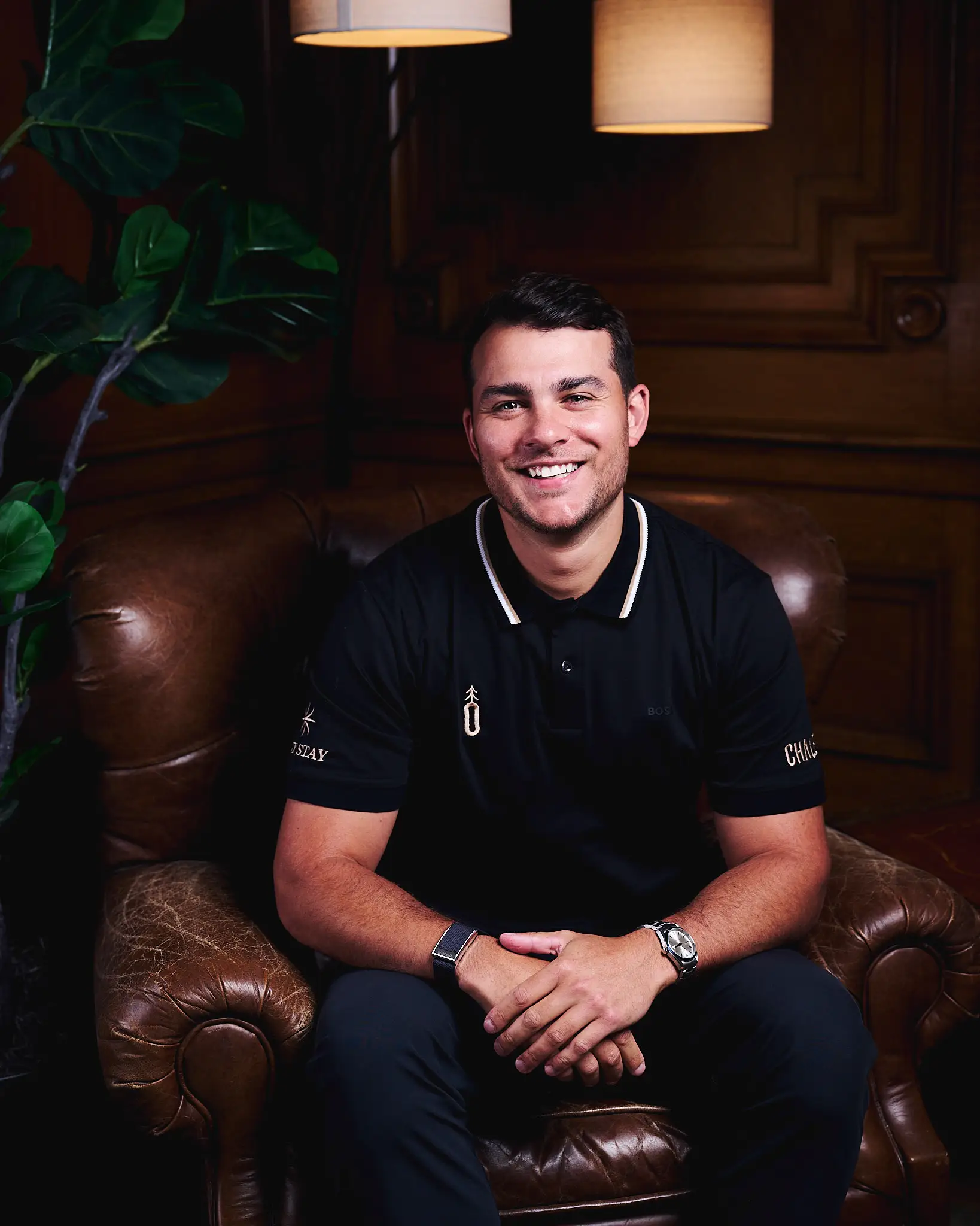 Smiling person in a black polo shirt sits in a brown leather armchair in a wood-paneled lounge.