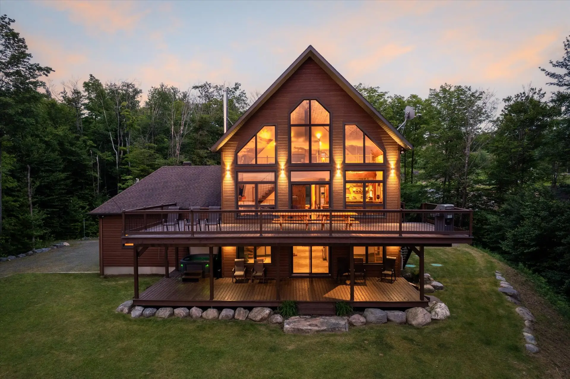 Illuminated A-frame wood cabin with large windows and decks sits in a wooded landscape at dusk.
