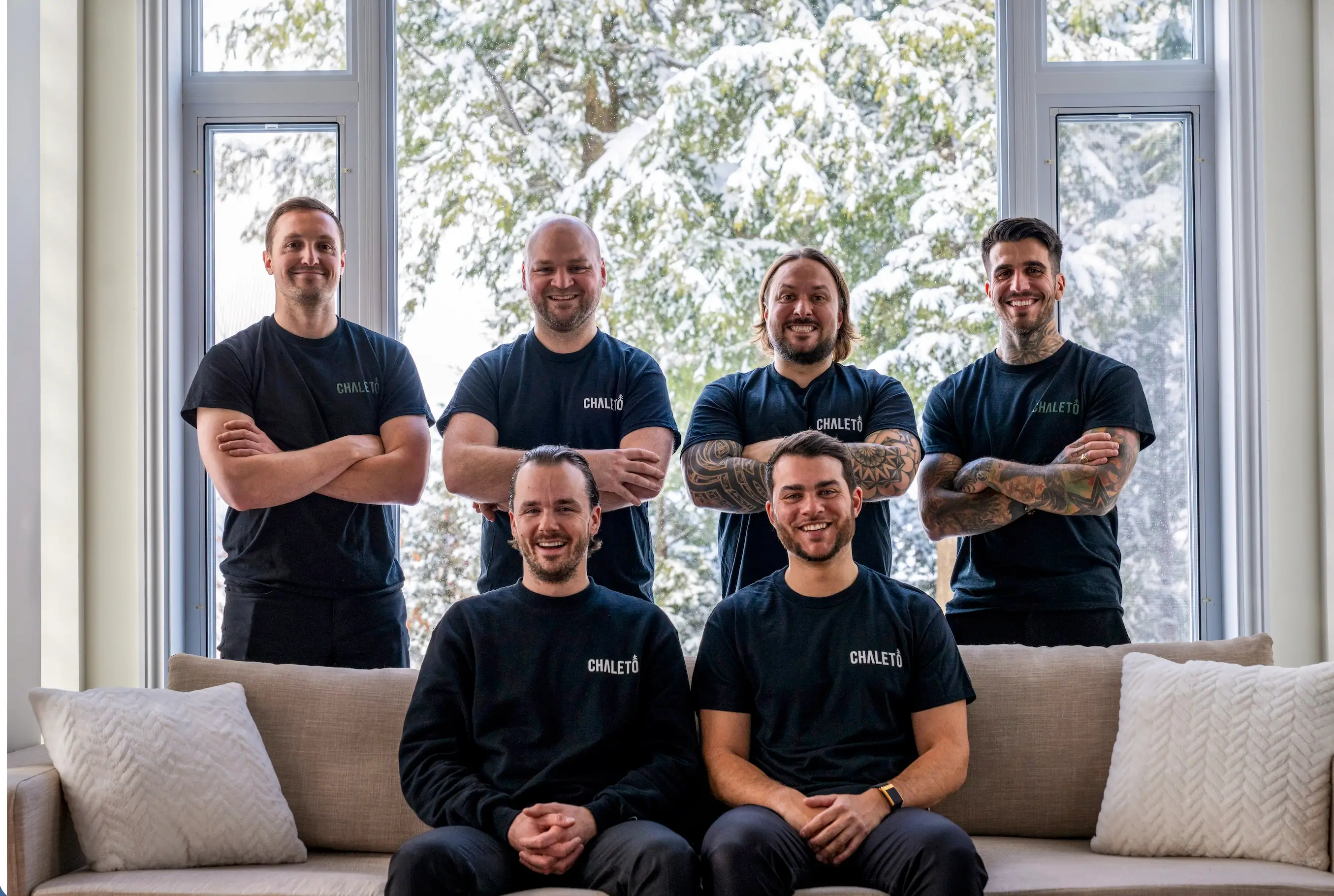Six people in matching CHALETÖ shirts pose indoors by large windows overlooking snowy trees.