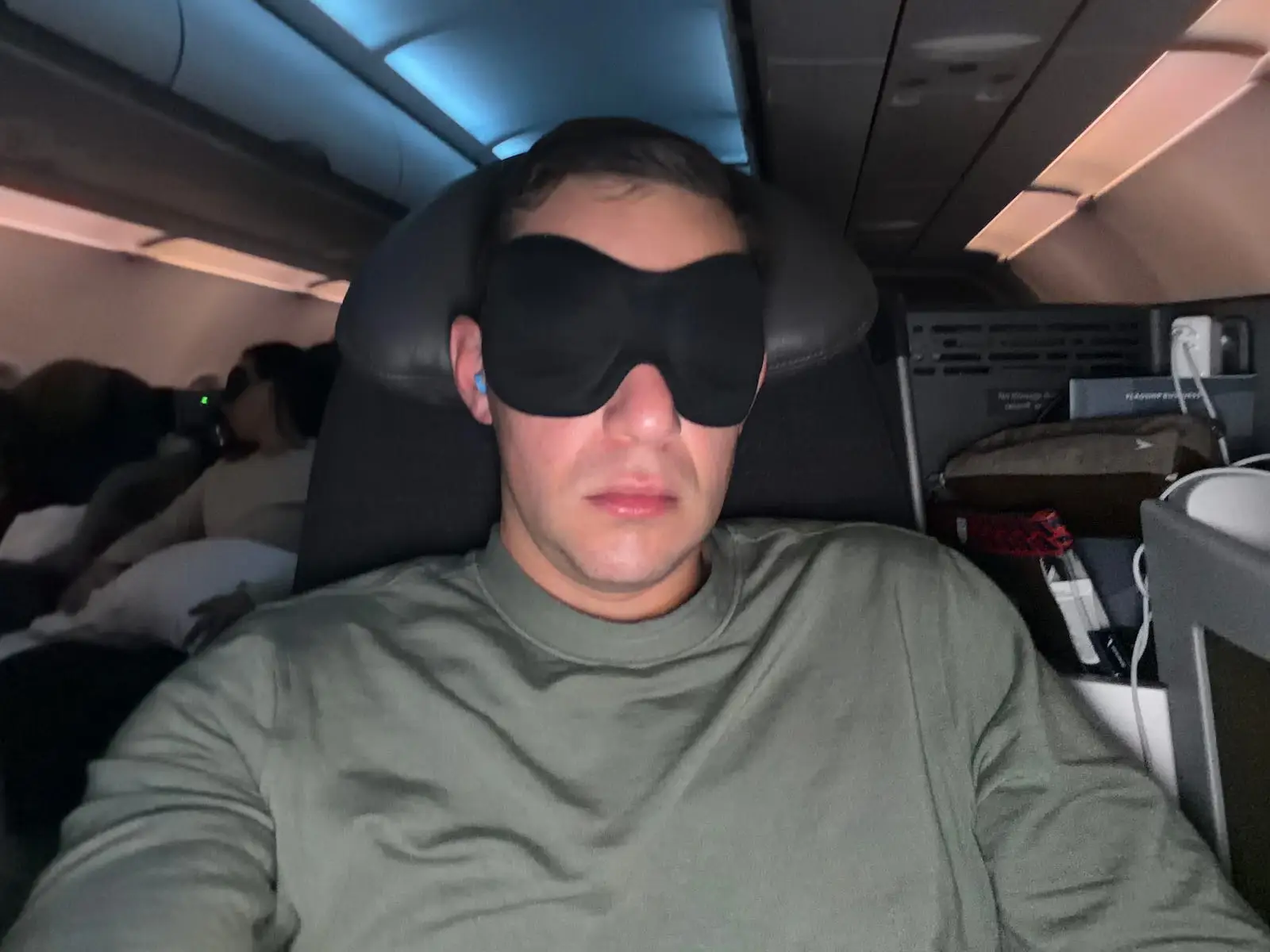 Passenger wearing a black sleep mask sits in a dimly lit airplane cabin with overhead lights.