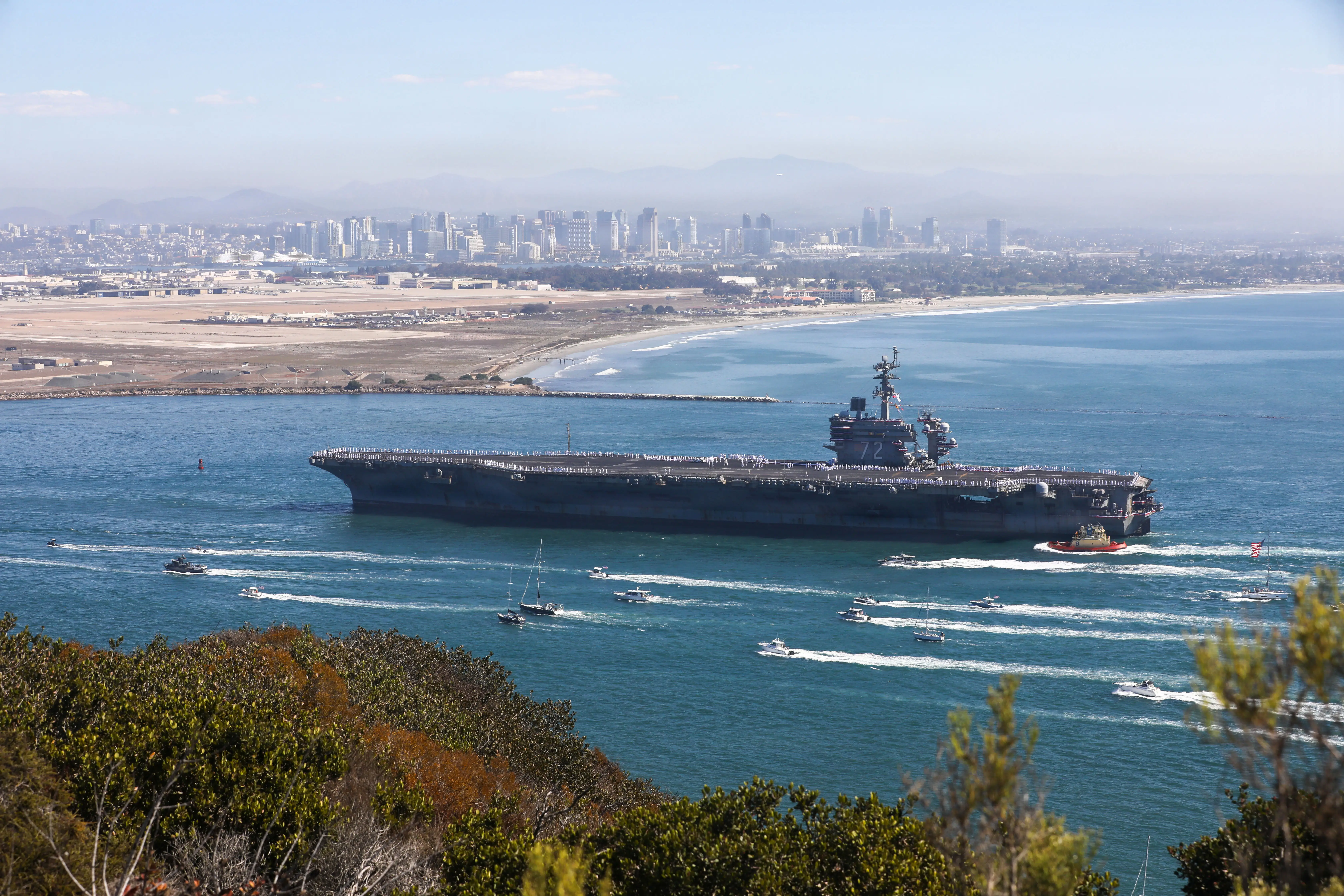 Aircraft carrier transits a coastal harbor with escort boats, shoreline, and a distant city skyline.