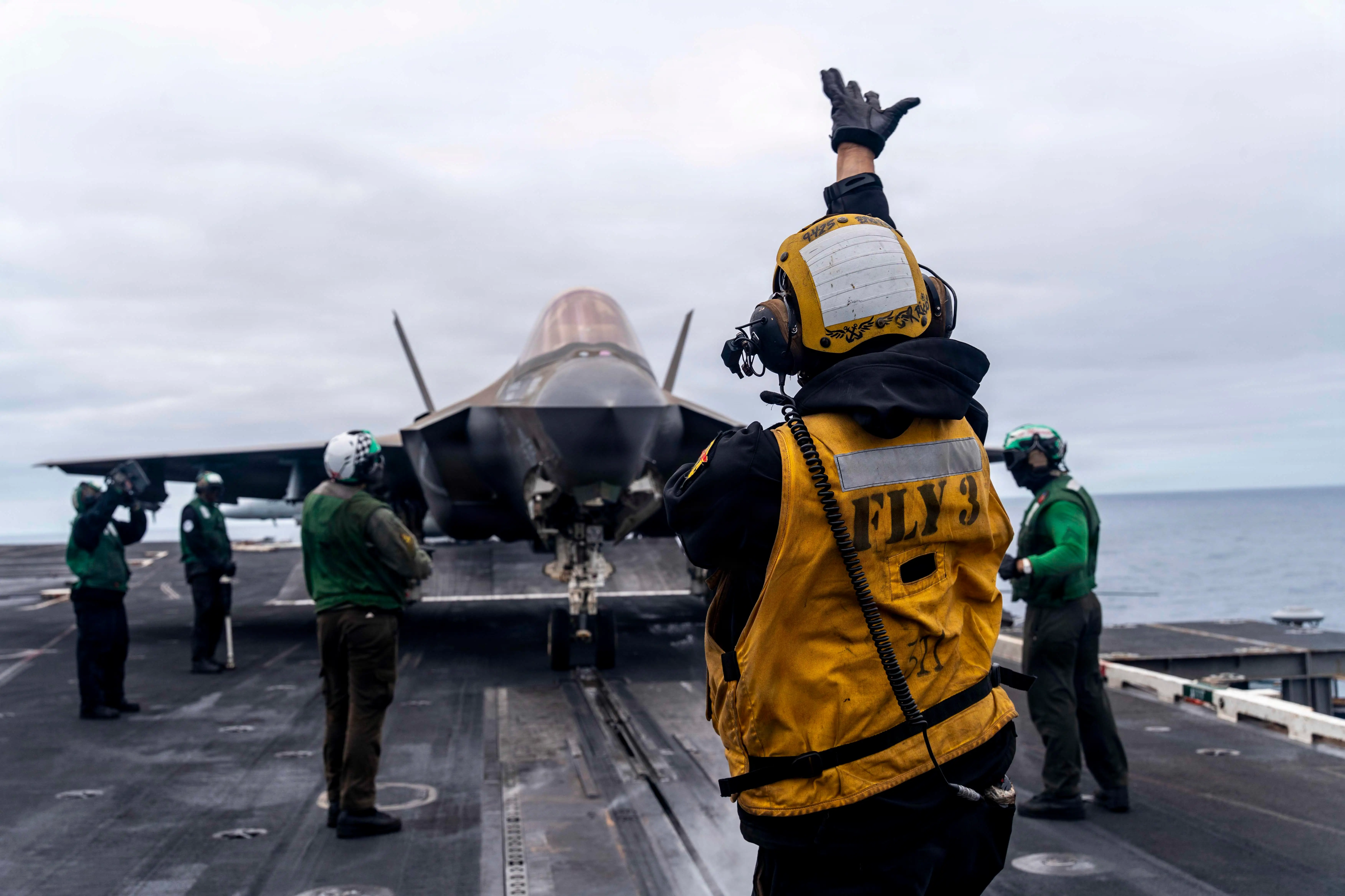 Flight deck crew signal near an F-35 fighter jet on an aircraft carrier at sea.