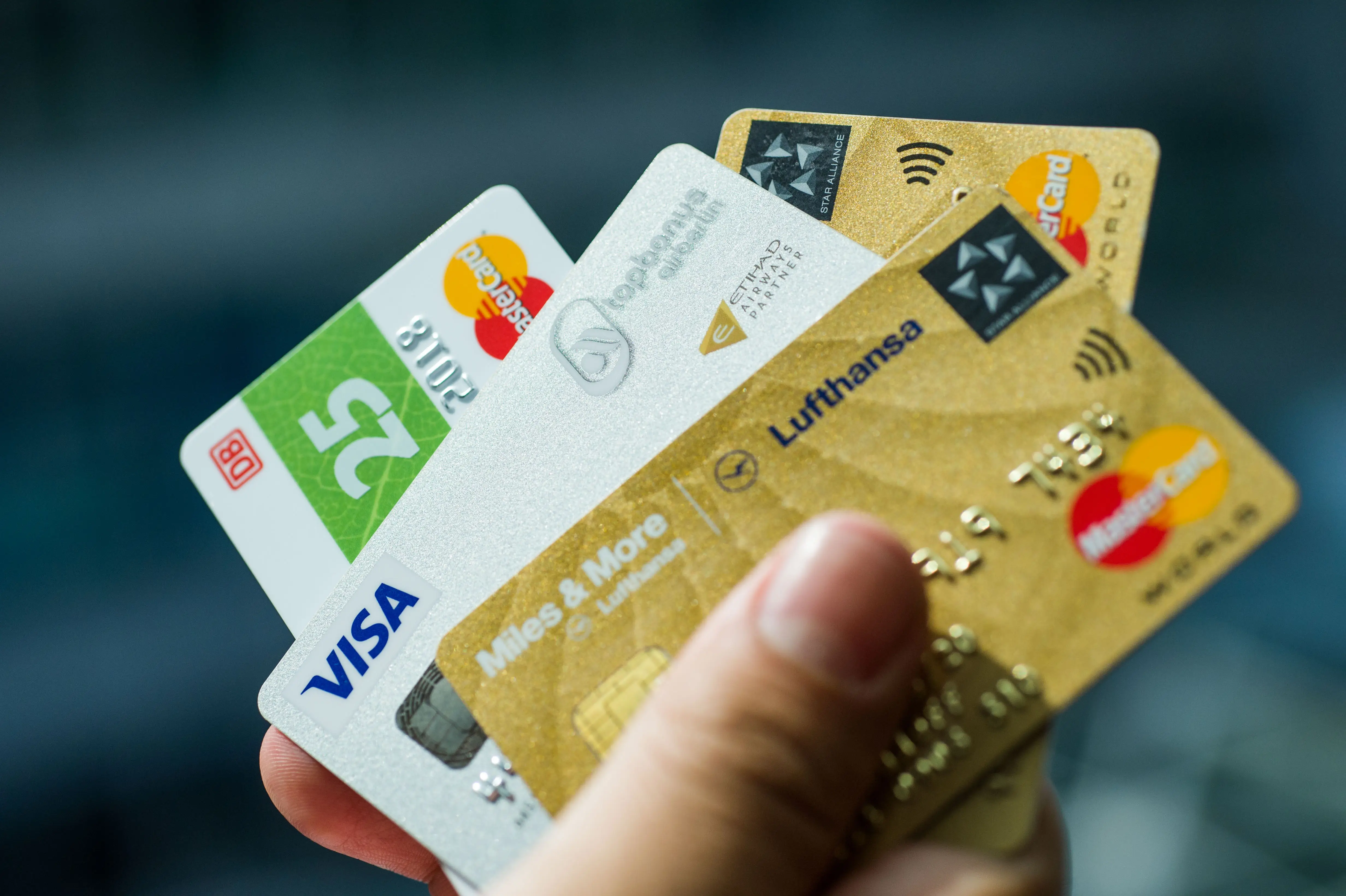 A hand holds several overlapping Visa and Mastercard payment cards, including Lufthansa Miles & More and Deutsche Bahn cards.