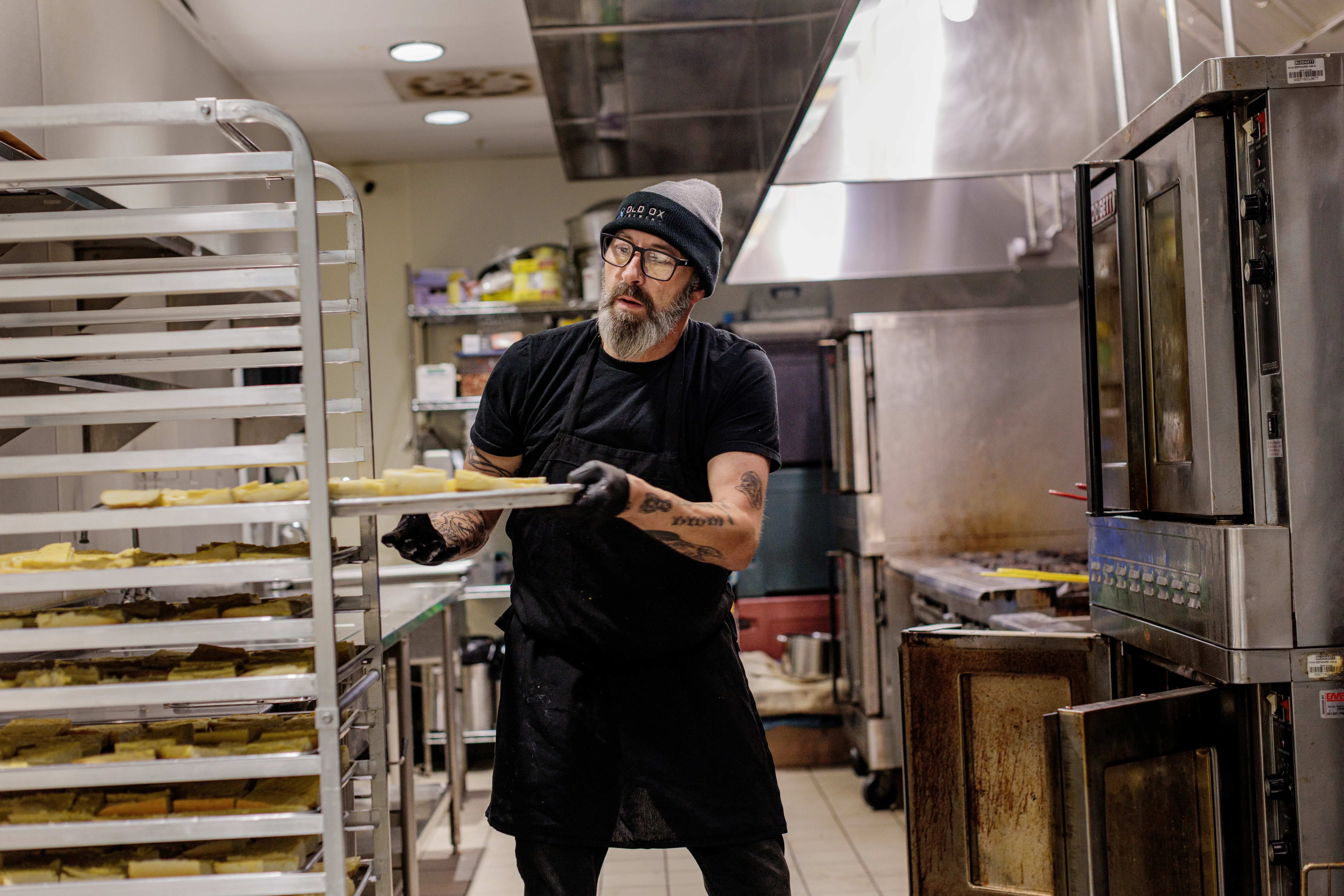 Kitchen worker in black clothing slides a tray of prepared food onto a rack beside commercial ovens.