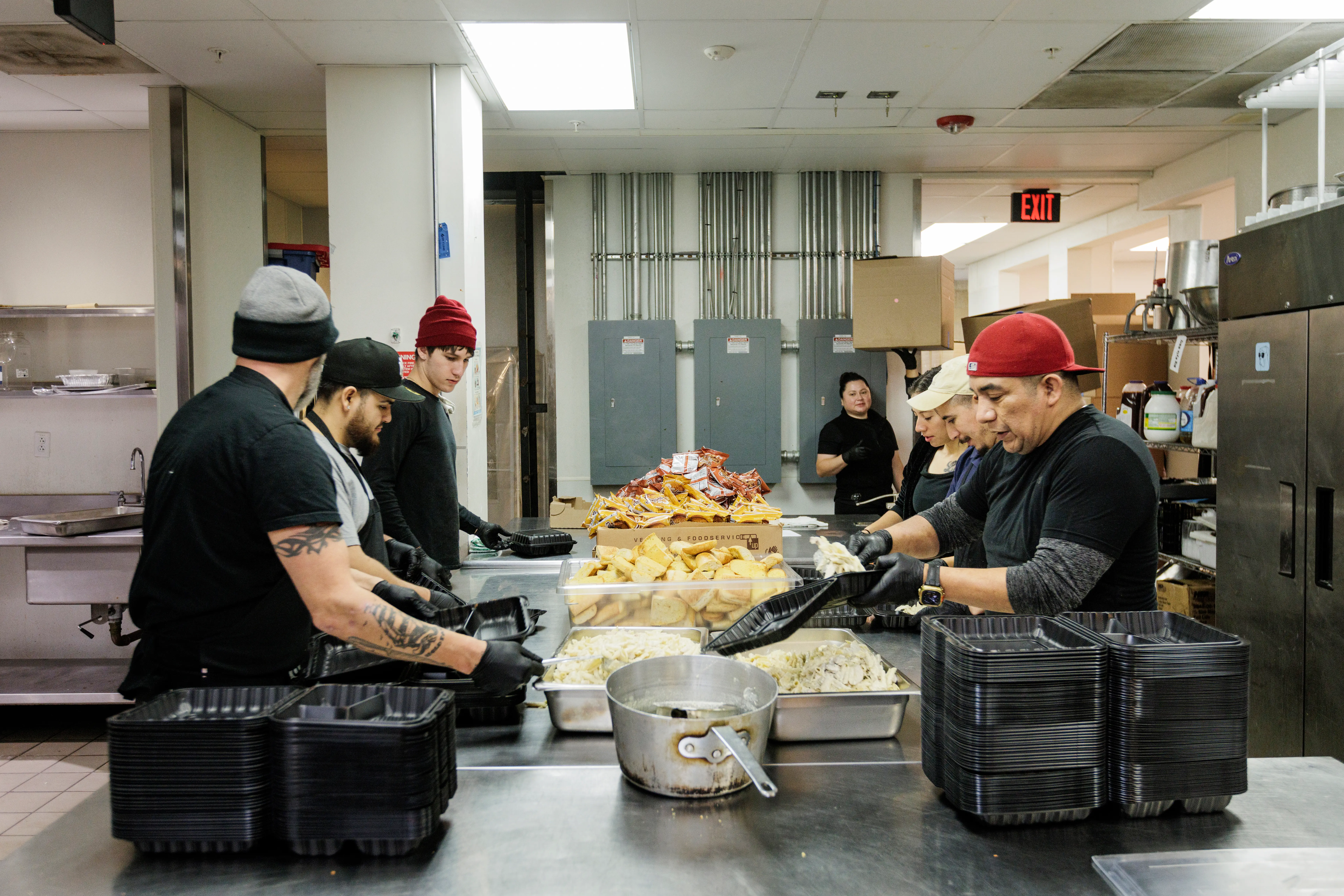 Kitchen workers pack prepared food into black containers around a stainless steel prep table.