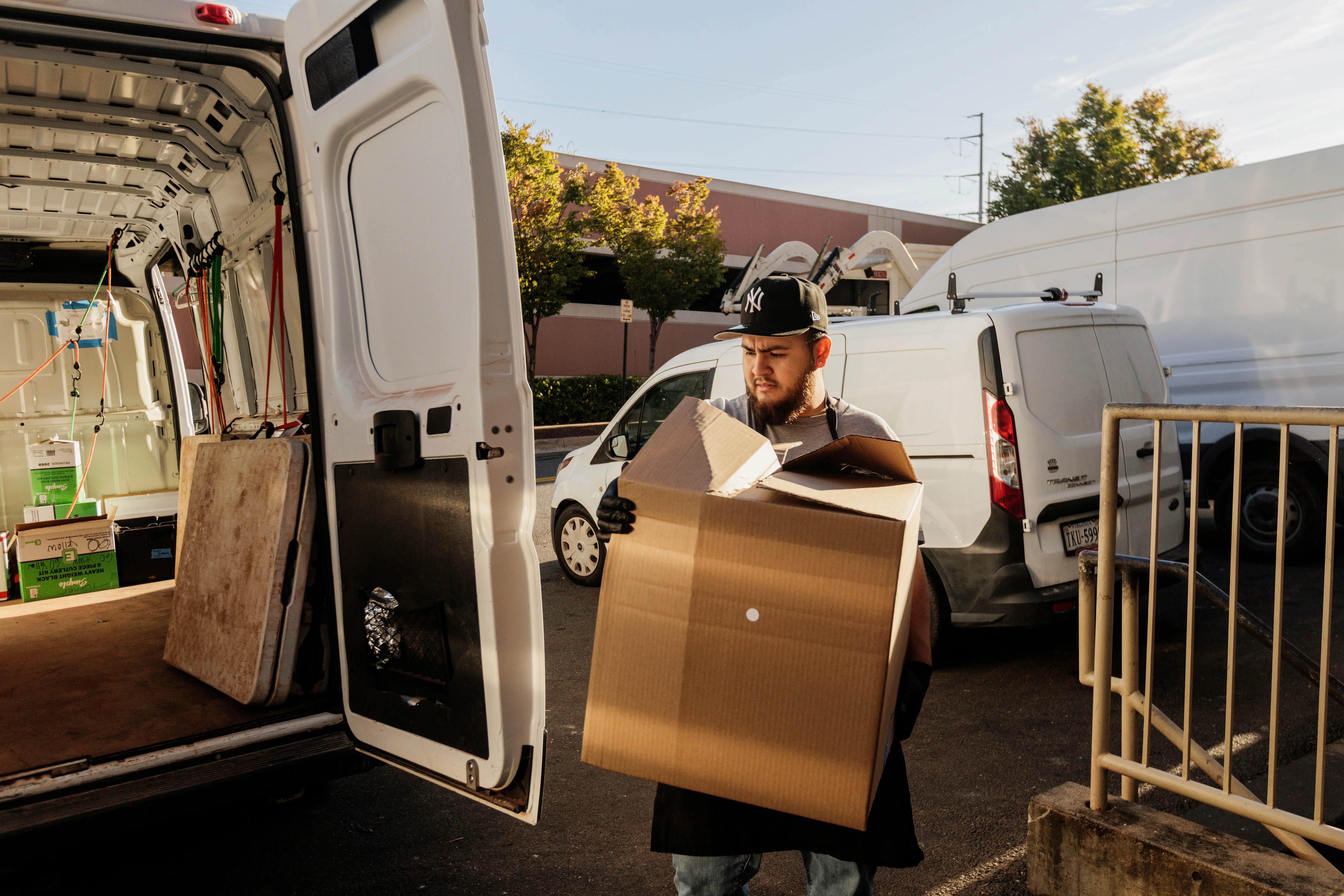 Person carries a large cardboard box beside open white cargo vans in a parking lot.