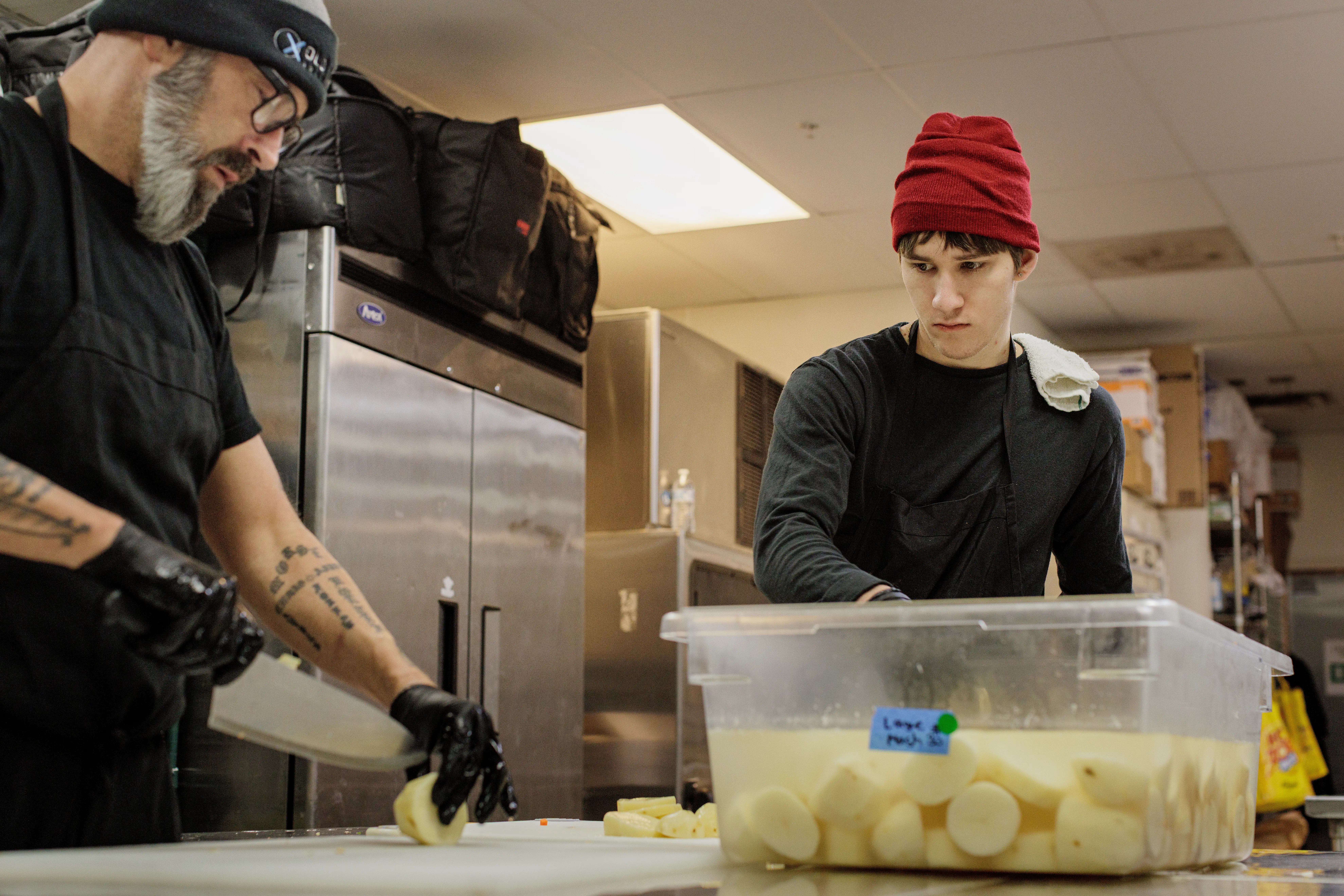 Two kitchen workers prepare peeled potatoes beside a labeled plastic tub in a commercial kitchen.