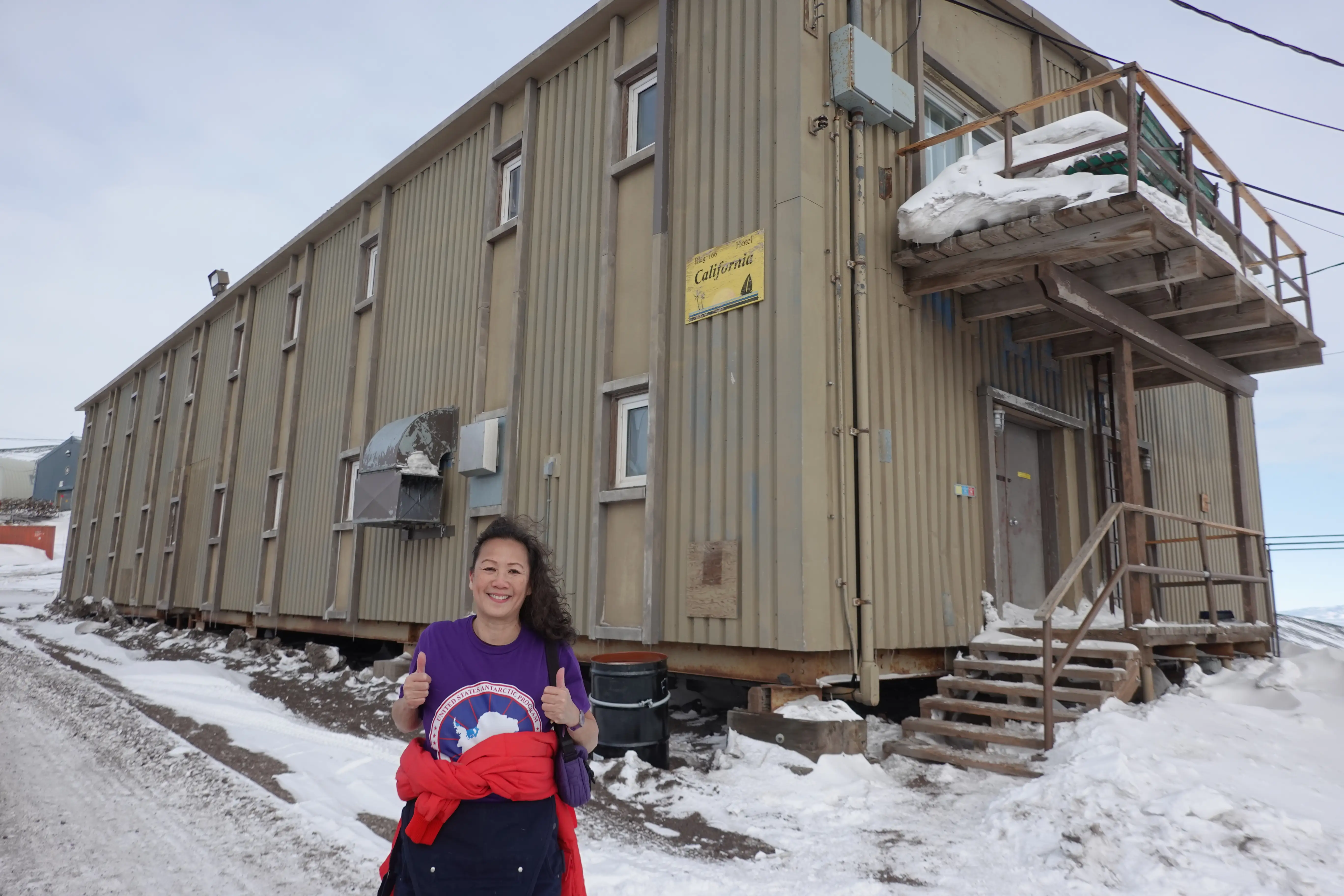 Person giving two thumbs up outside a beige building with snowy stairs and a yellow Hotel California sign.