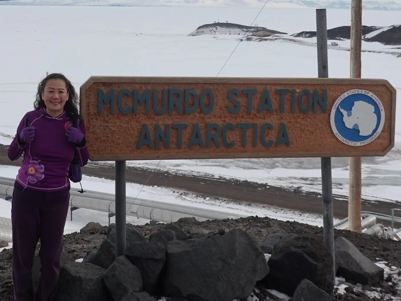 Person standing beside a large McMurdo Station Antarctica sign in a snowy Antarctic landscape.