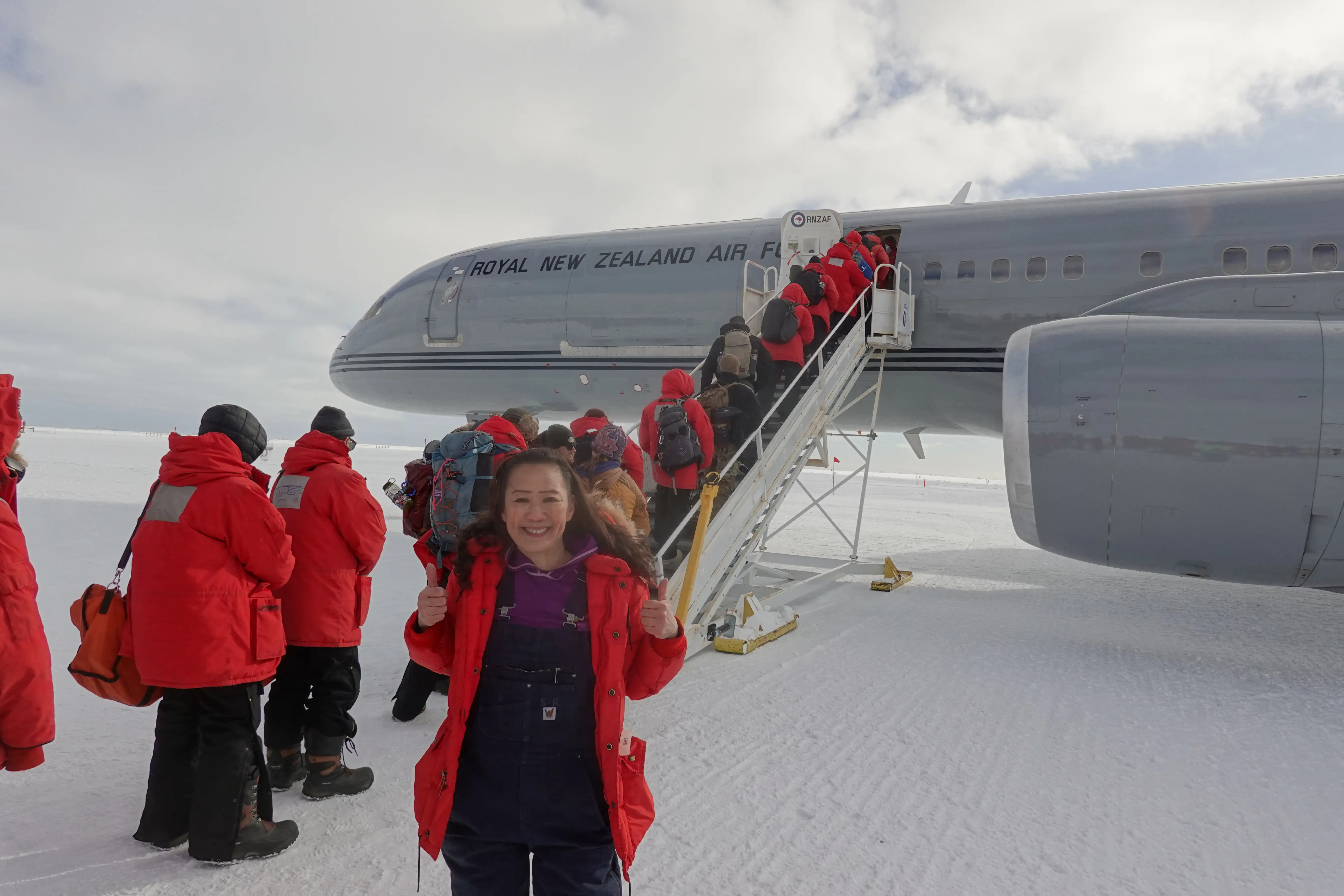 Travelers in red parkas board a Royal New Zealand Air Force aircraft on a snowy airfield.