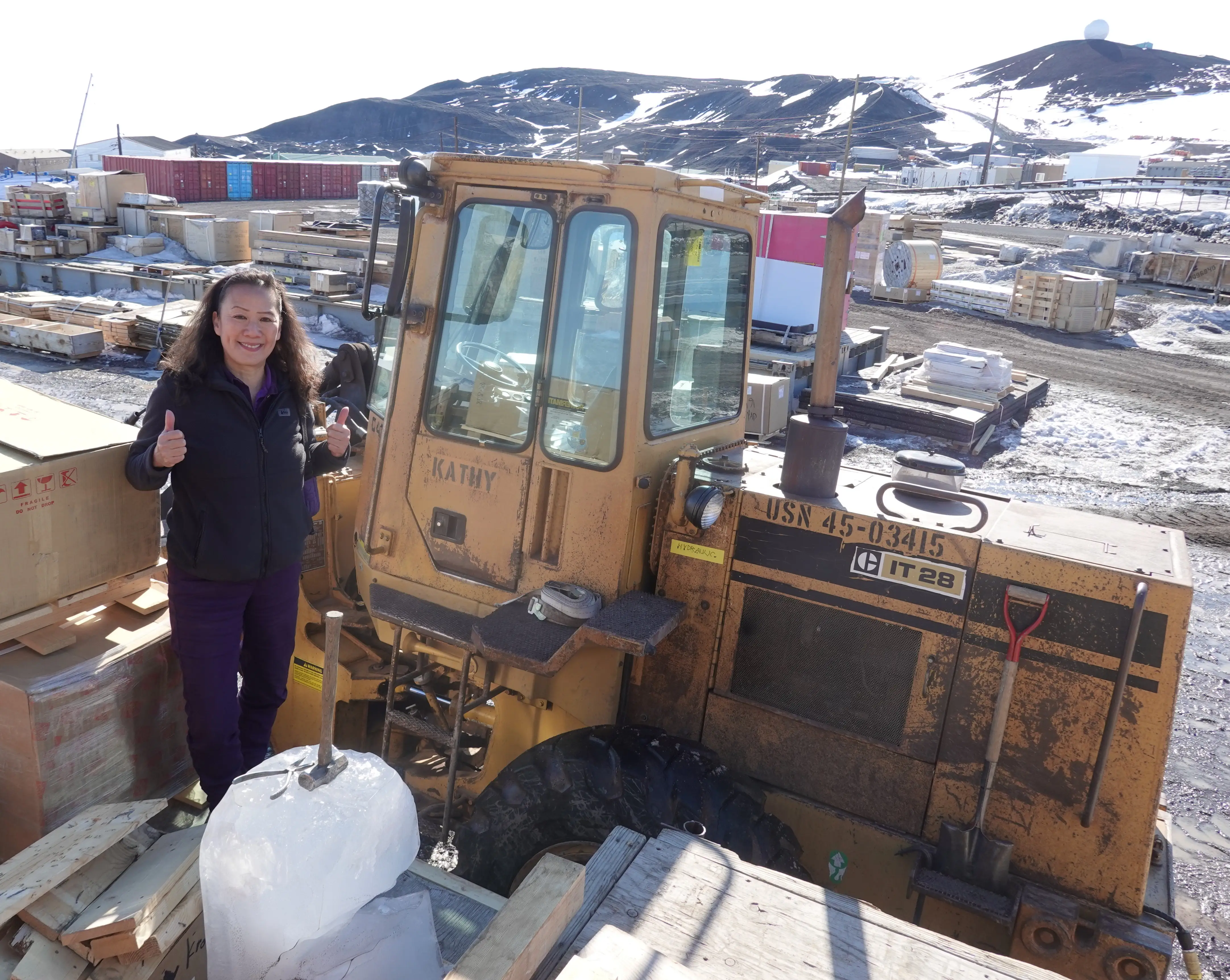 Person giving thumbs-up beside a muddy CAT IT 28 loader in a snowy industrial yard with mountains behind.