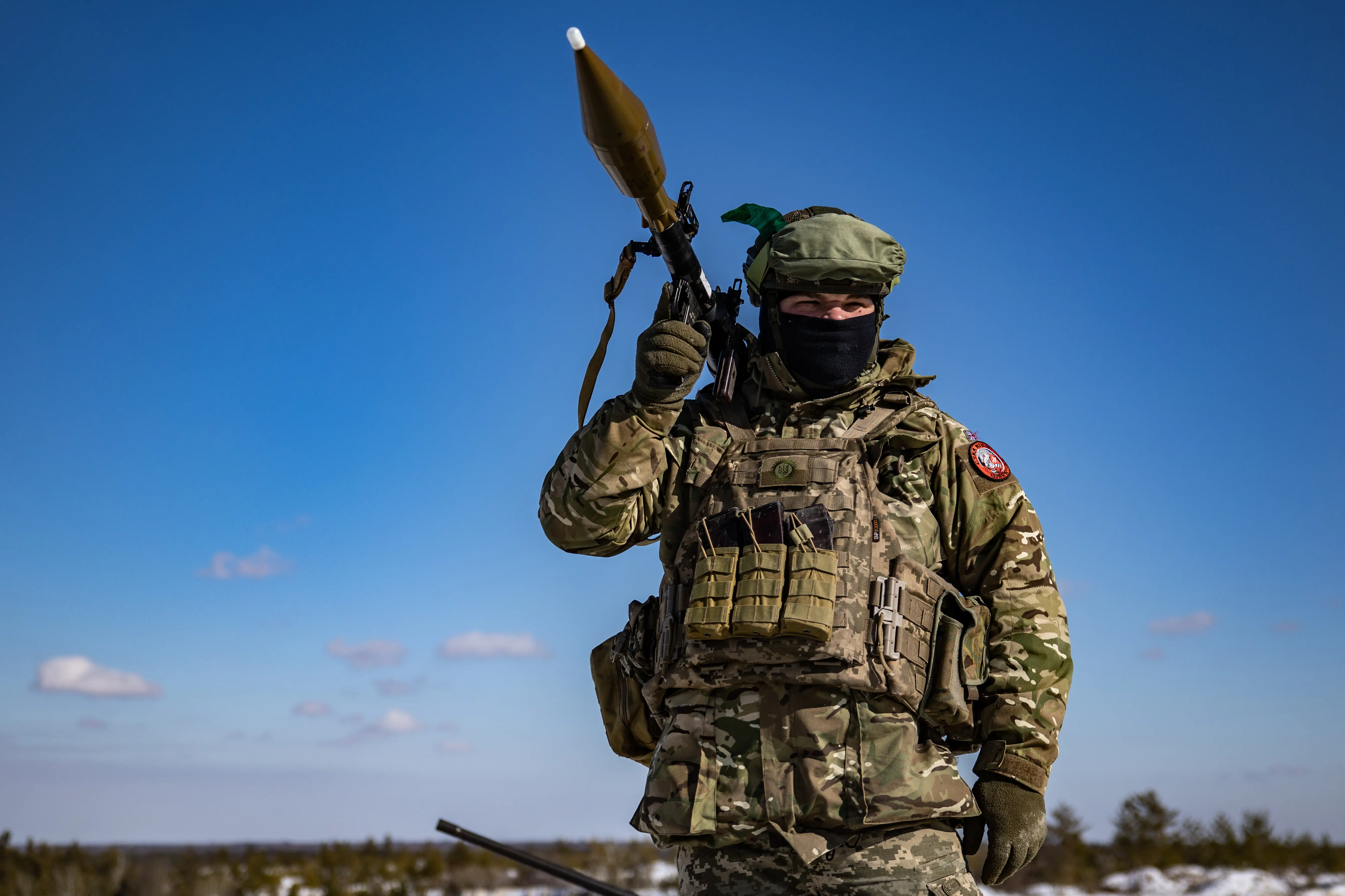 Masked person in camouflage gear carries a rocket-propelled grenade launcher in a snowy open landscape under a blue sky.