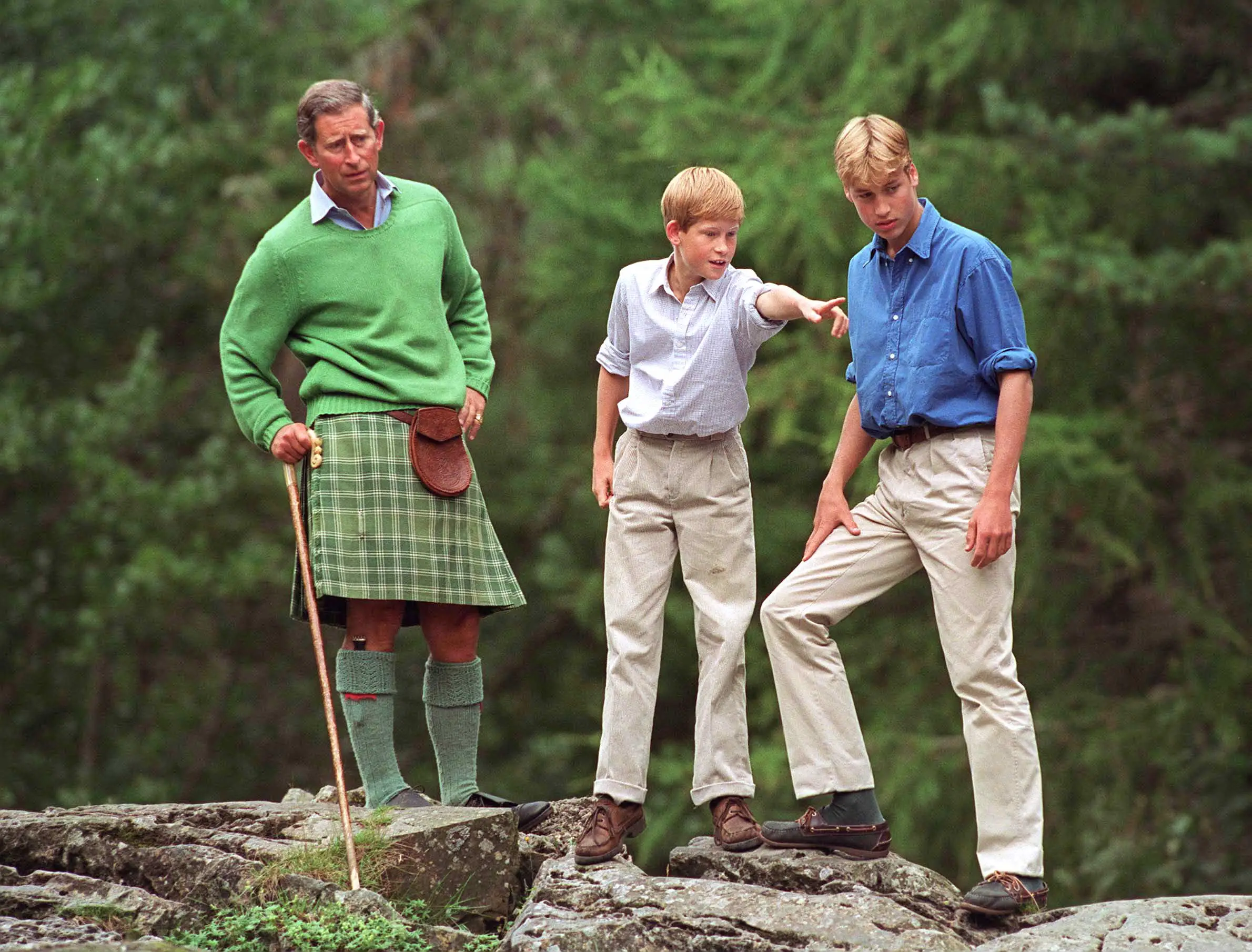 King Charles III, Prince William, and Prince Harry stand on rocks in a wooded outdoor setting.