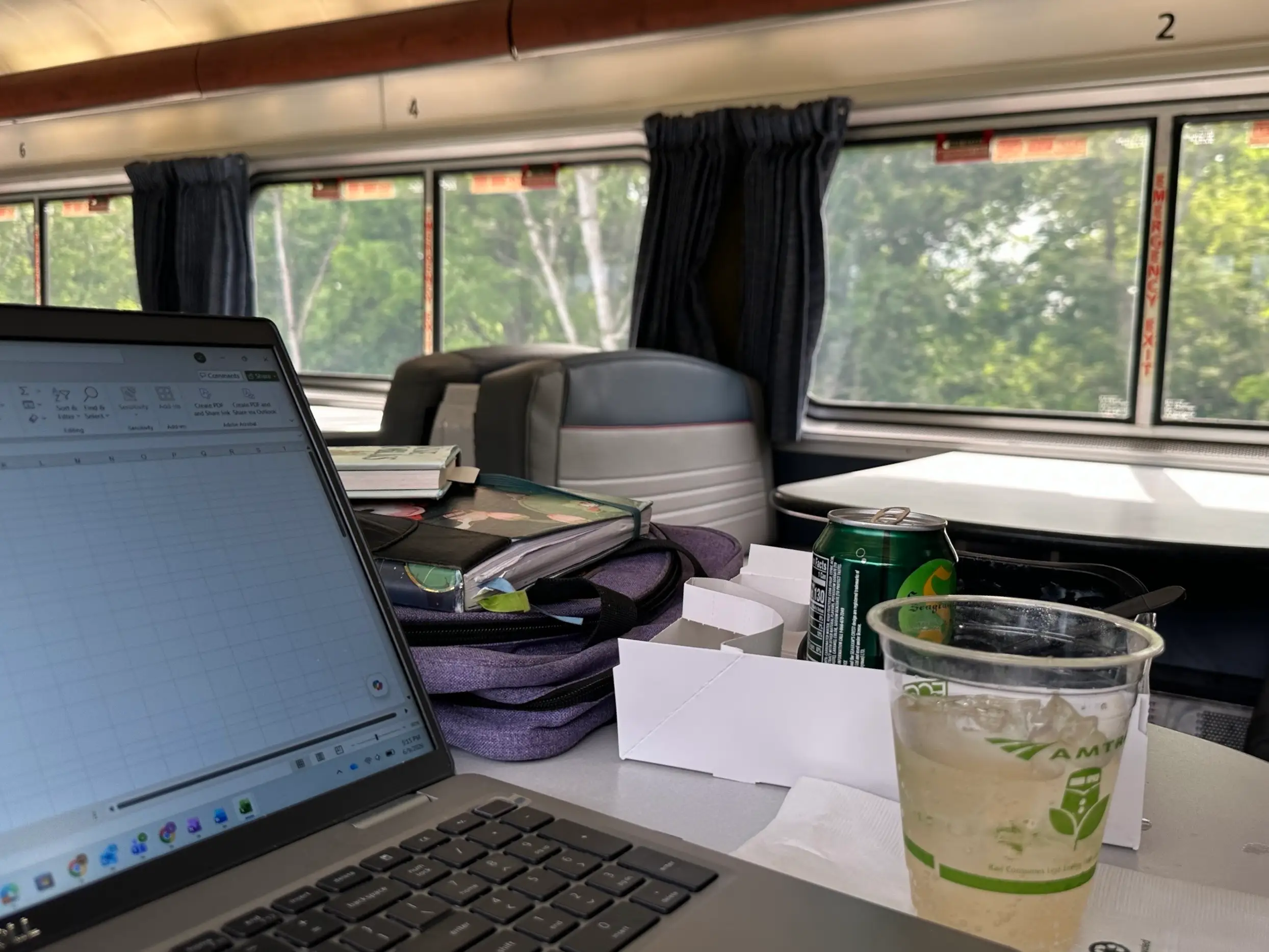The writer's laptop with a spreadsheet sits on a passenger train table beside drinks, boxes, bags, seats, and curtained windows.