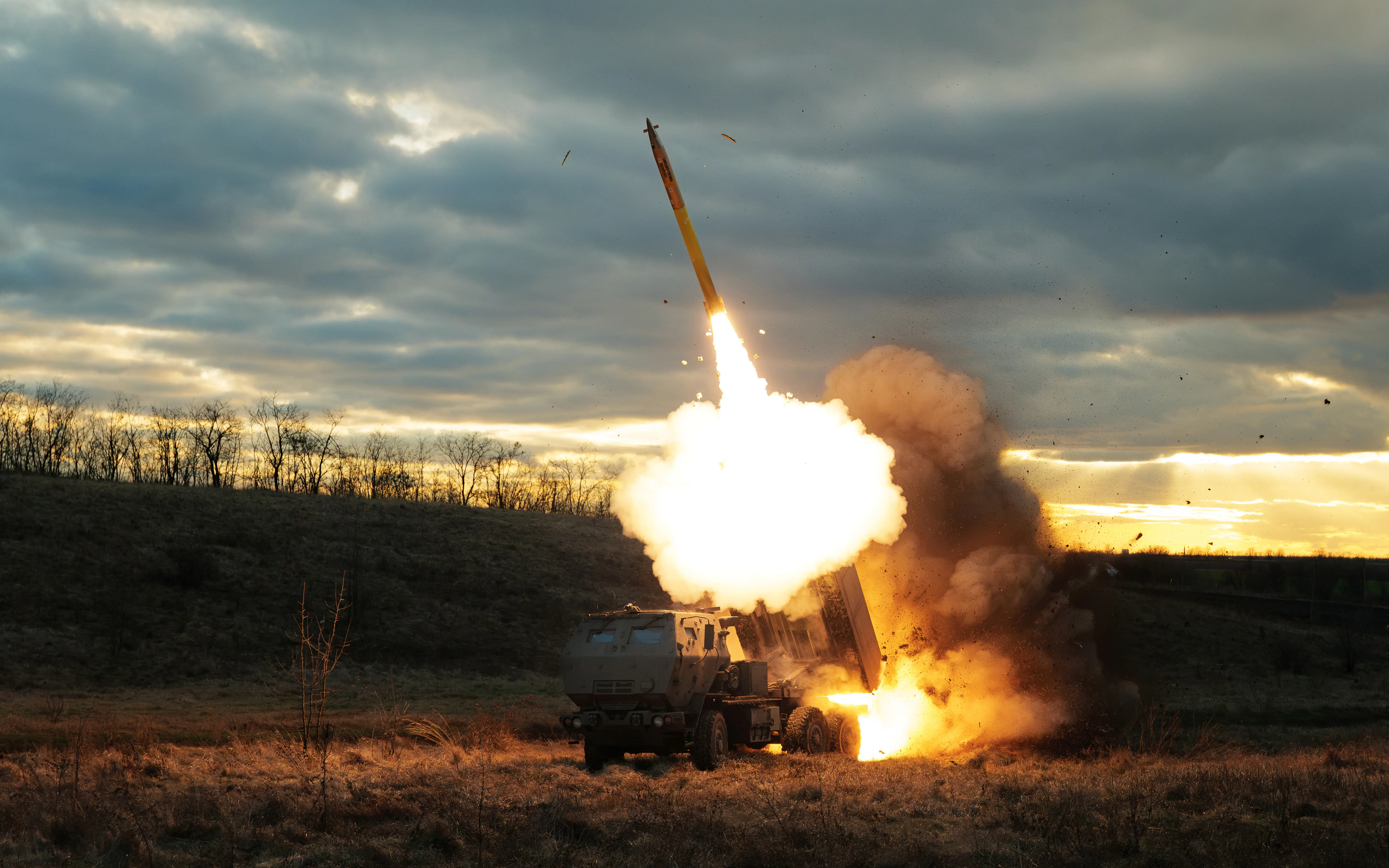 Truck-mounted rocket launcher fires a missile from a grassy field under a cloudy sunset sky.