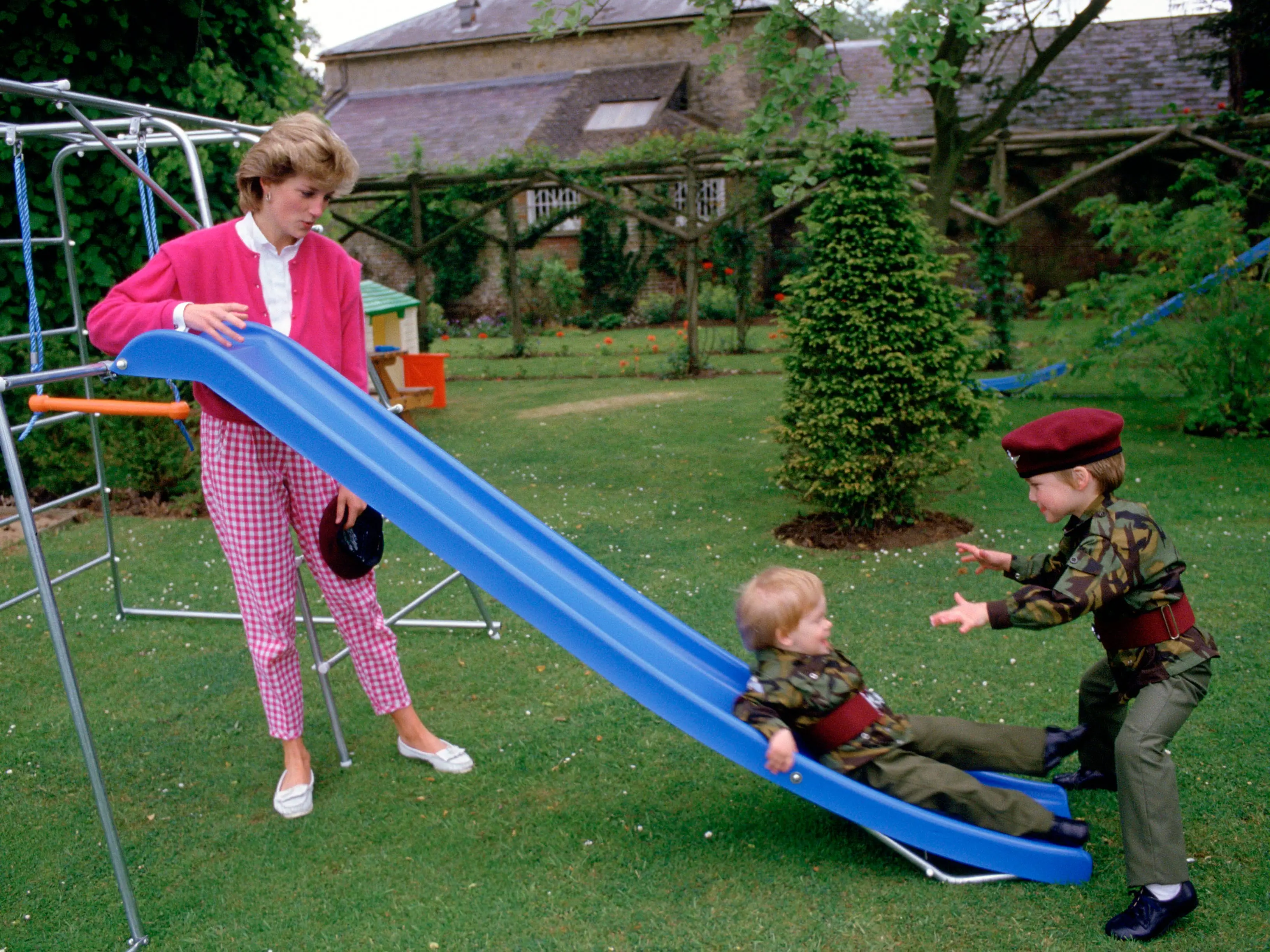 Princess Diana stands beside a blue garden slide while two children play on the lawn.