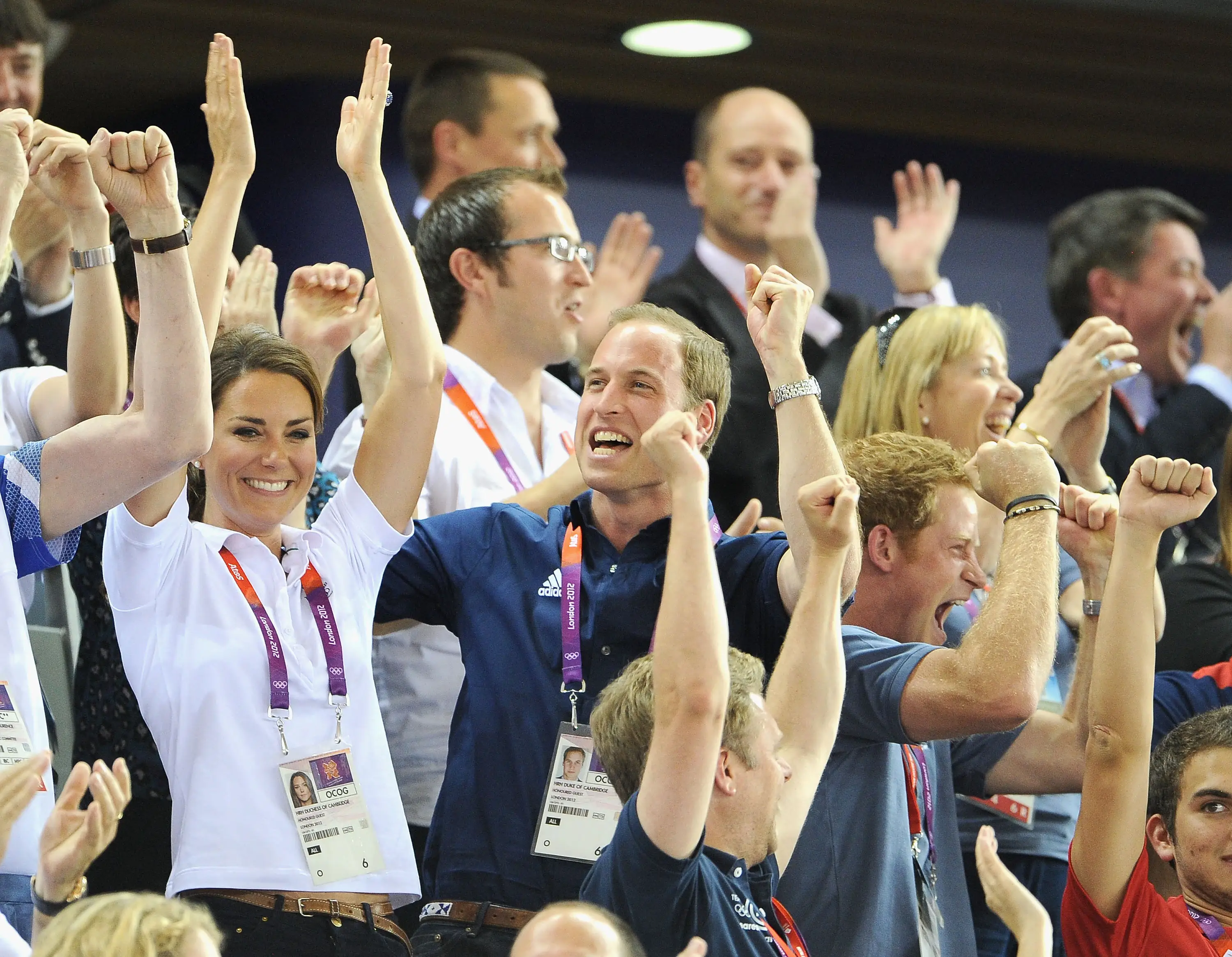 Prince William, Catherine, and Prince Harry cheer with raised arms among spectators at a sporting event.