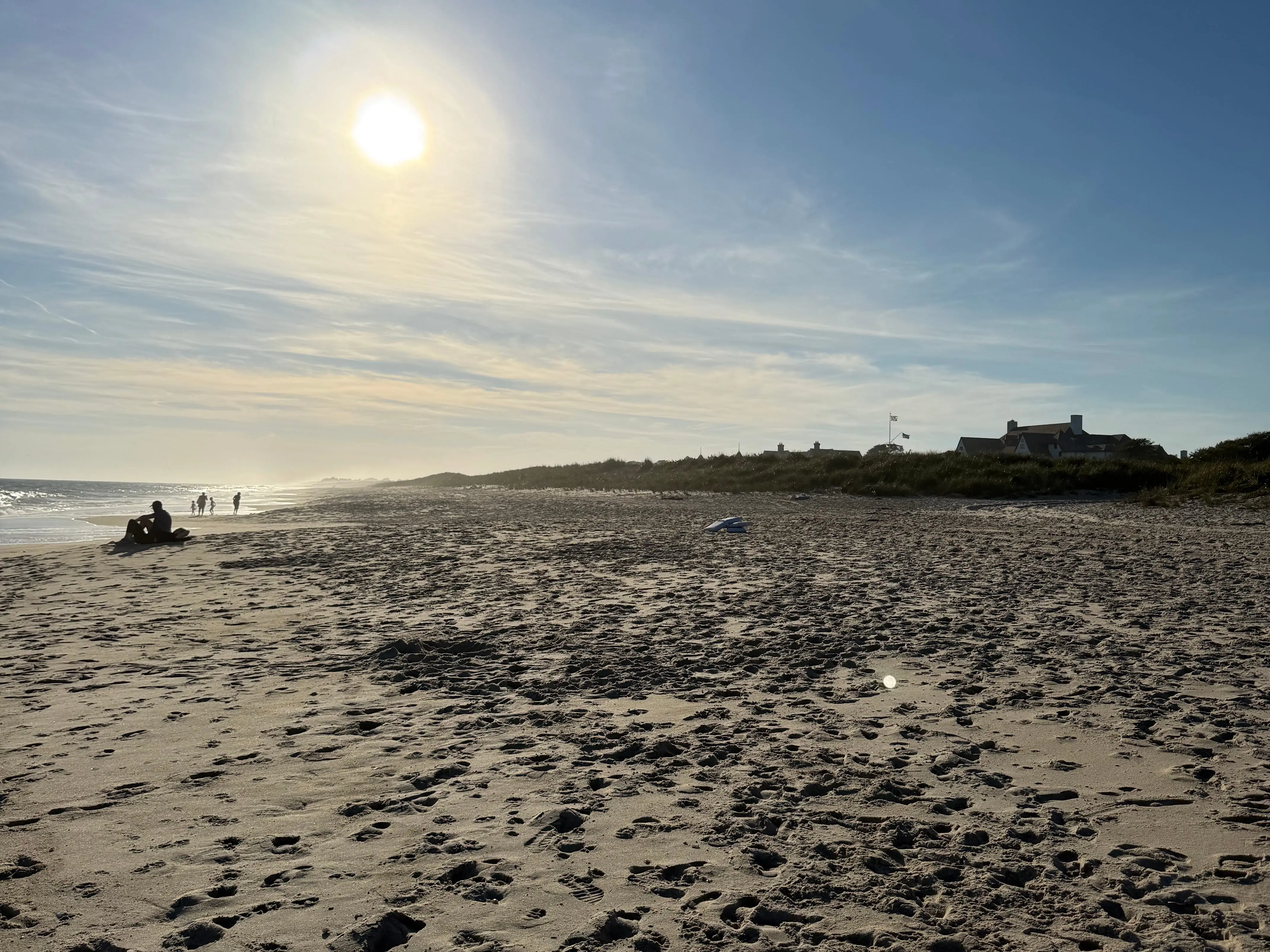 Low sun shines over a sandy beach in the Hamptons with footprints, ocean surf, distant people, dunes, and shoreline buildings.