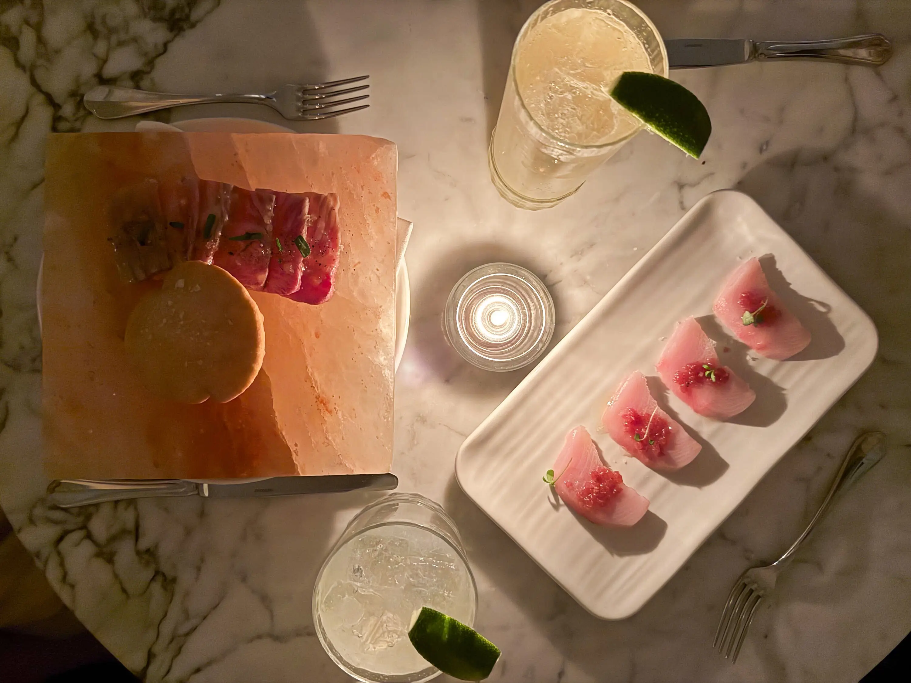 Overhead restaurant table with sliced fish dishes, lime-garnished drinks, a candle, and utensils on marble.