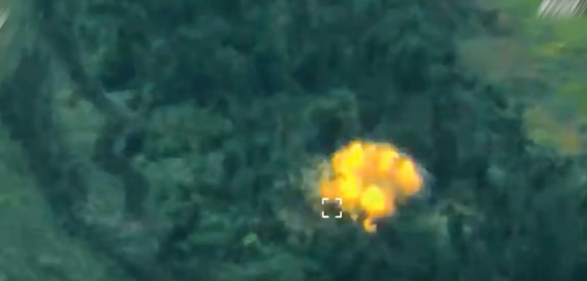 Thermal aerial view shows a bright explosion or fireball in a wooded area with a targeting reticle nearby.