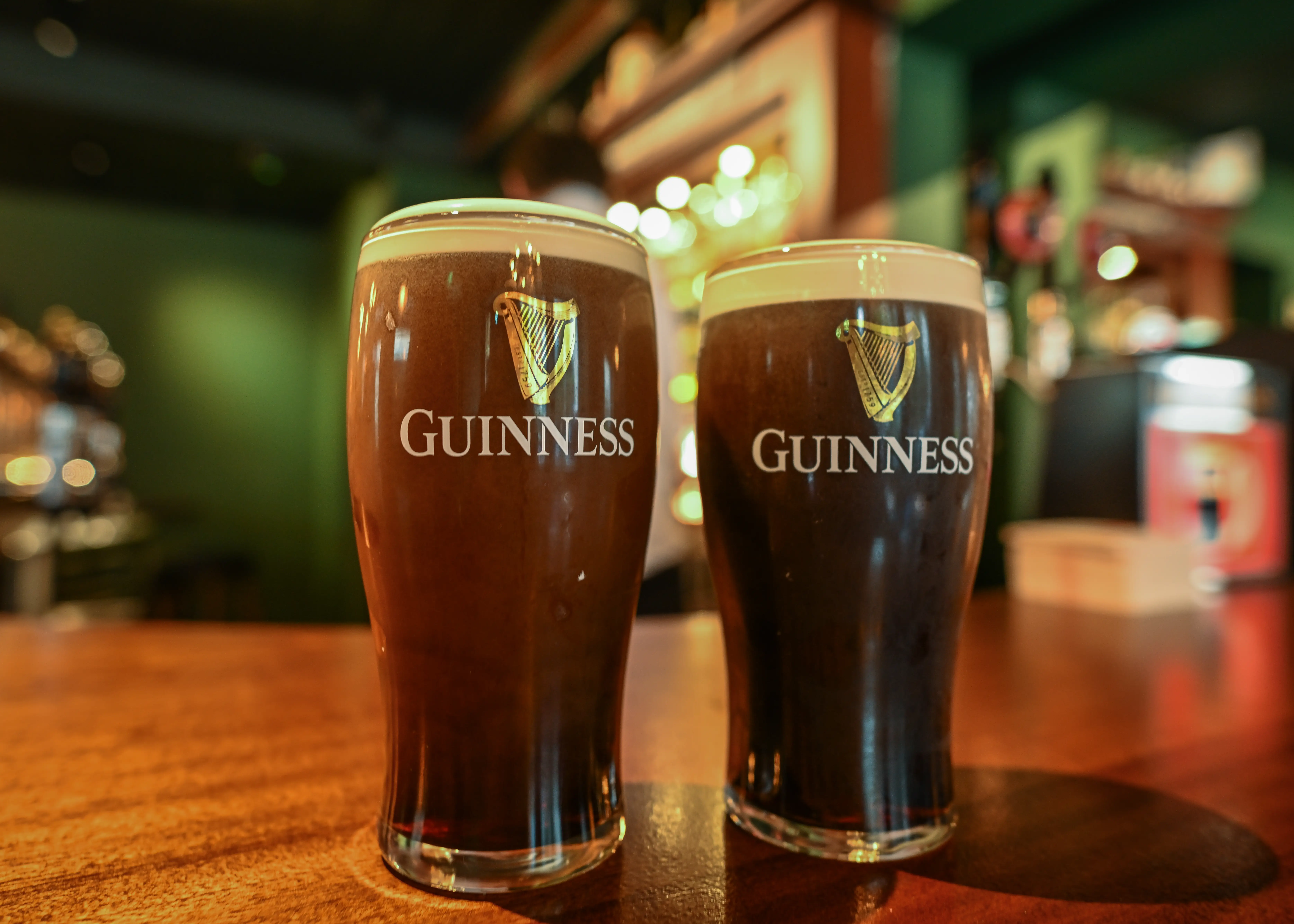 Two pints of GUINNESS stout sit on a wooden bar in a softly lit pub.