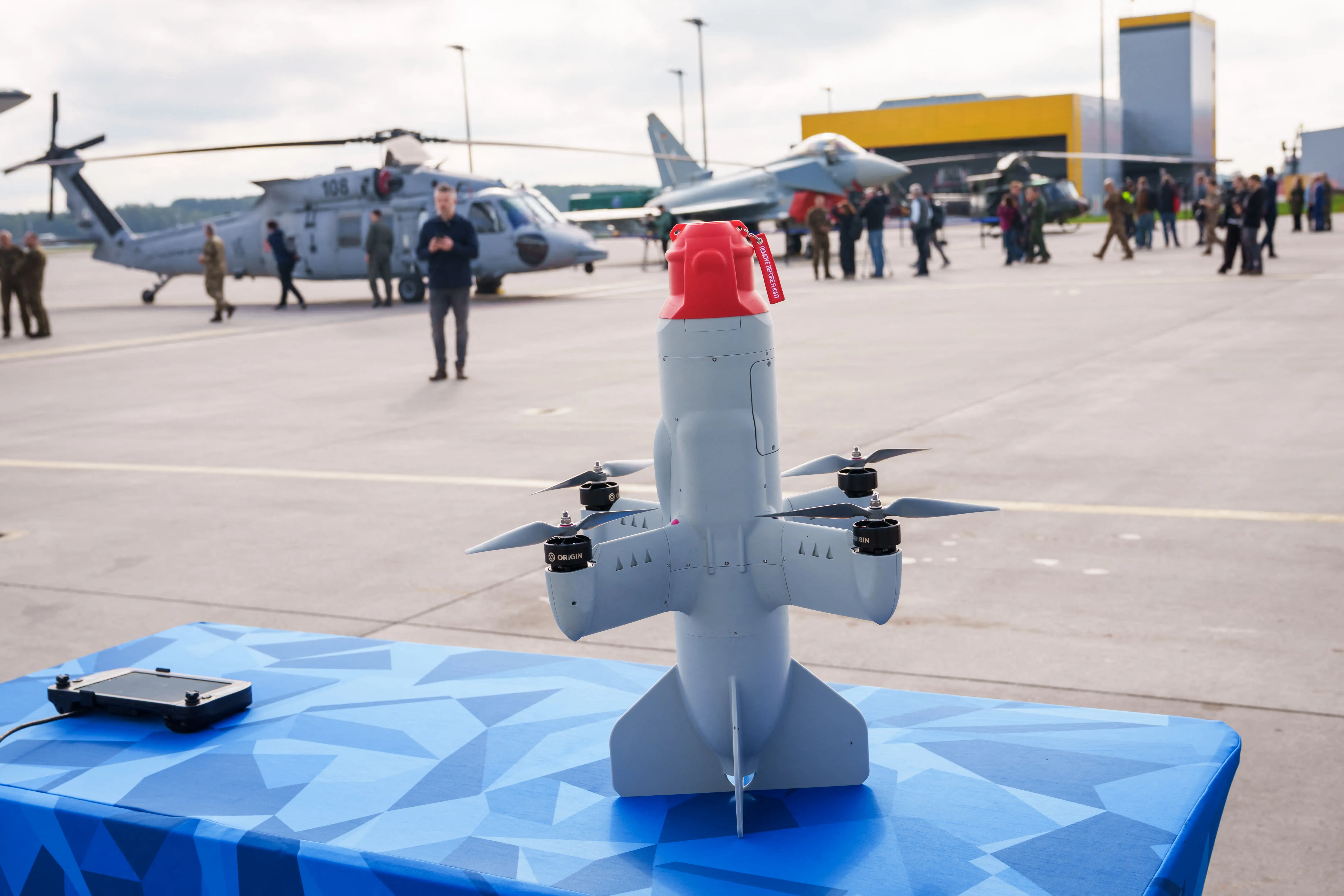 A small VTOL drone sits on a blue display table at an airfield with aircraft and visitors behind it.