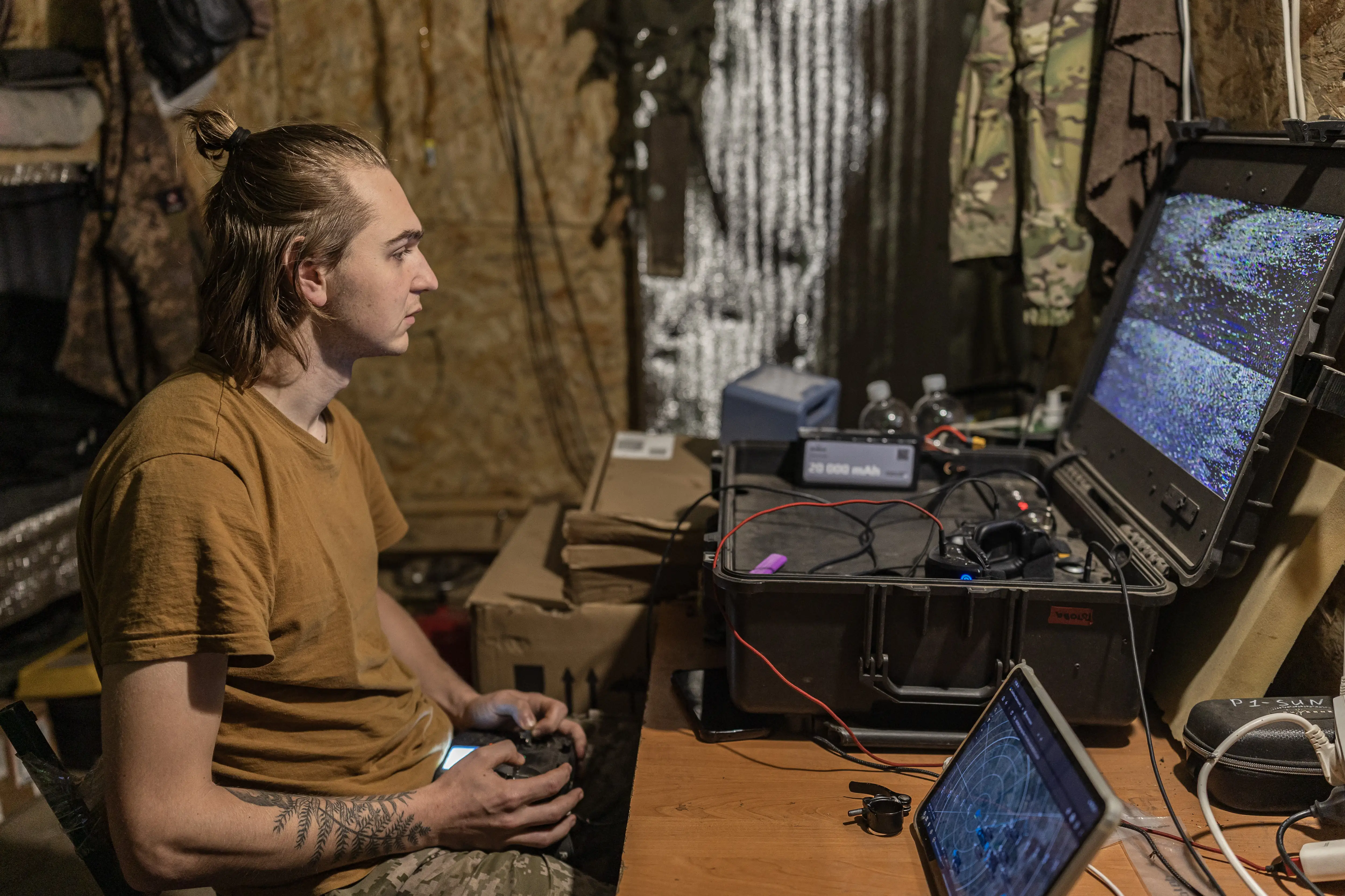 Person in camouflage pants operates a handheld controller beside rugged screens and electronics in a dim improvised workspace.