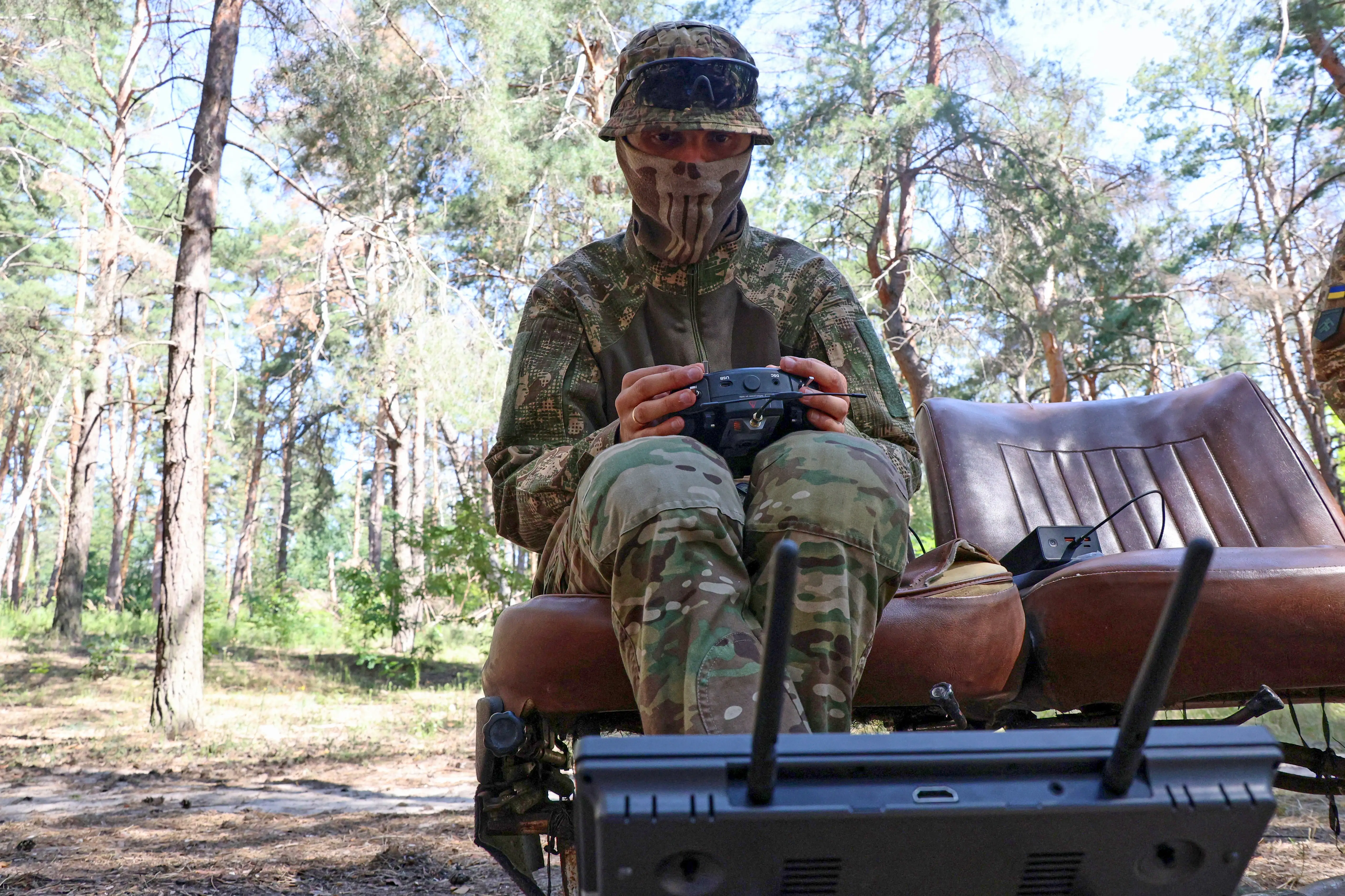 Camouflaged masked person sits in a forest operating a handheld remote control beside radio equipment.