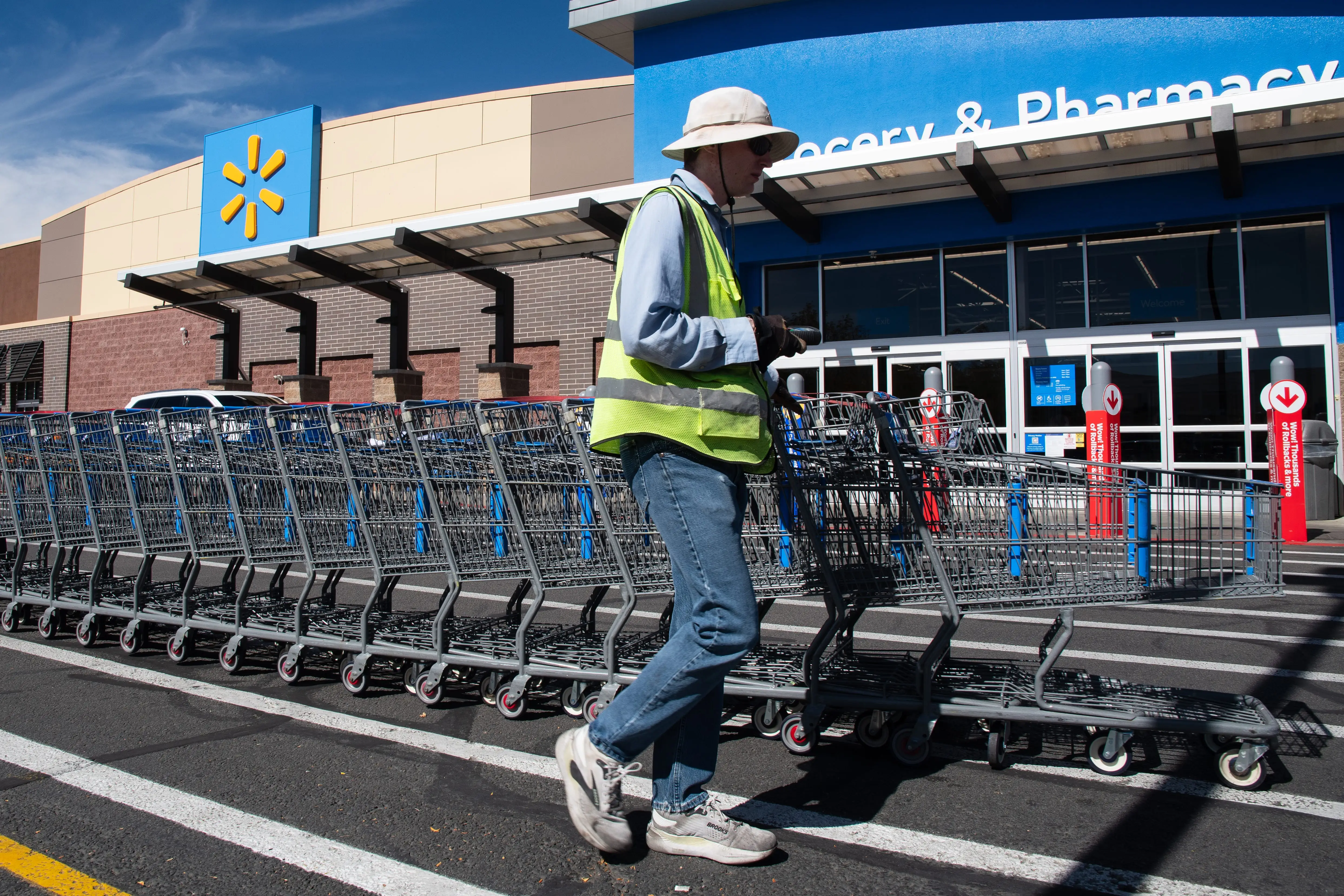 Worker in a reflective vest pushes rows of shopping carts outside a Walmart store entrance.