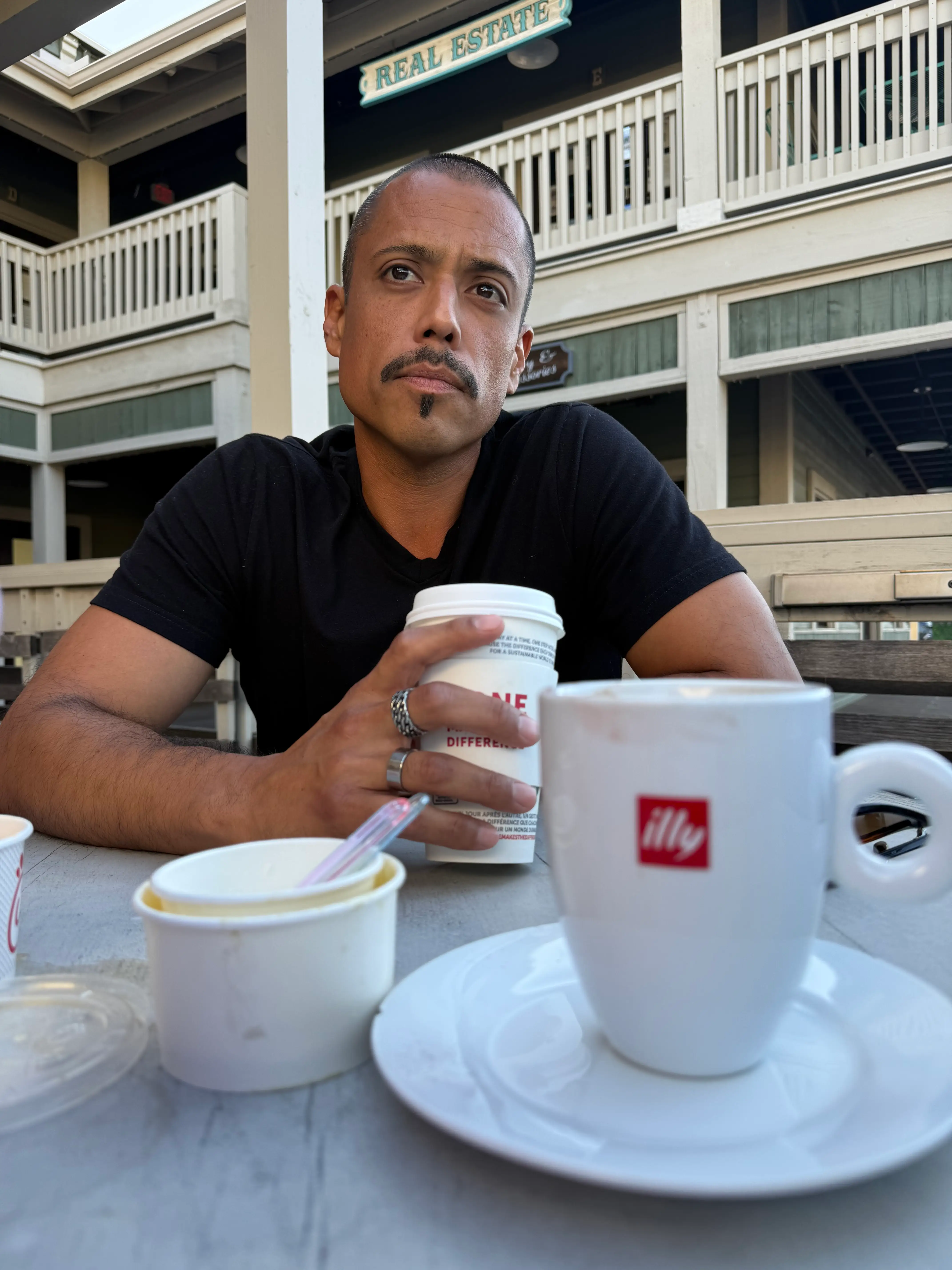 Person in a black shirt sits at an outdoor cafe table holding a paper cup near an illy mug.