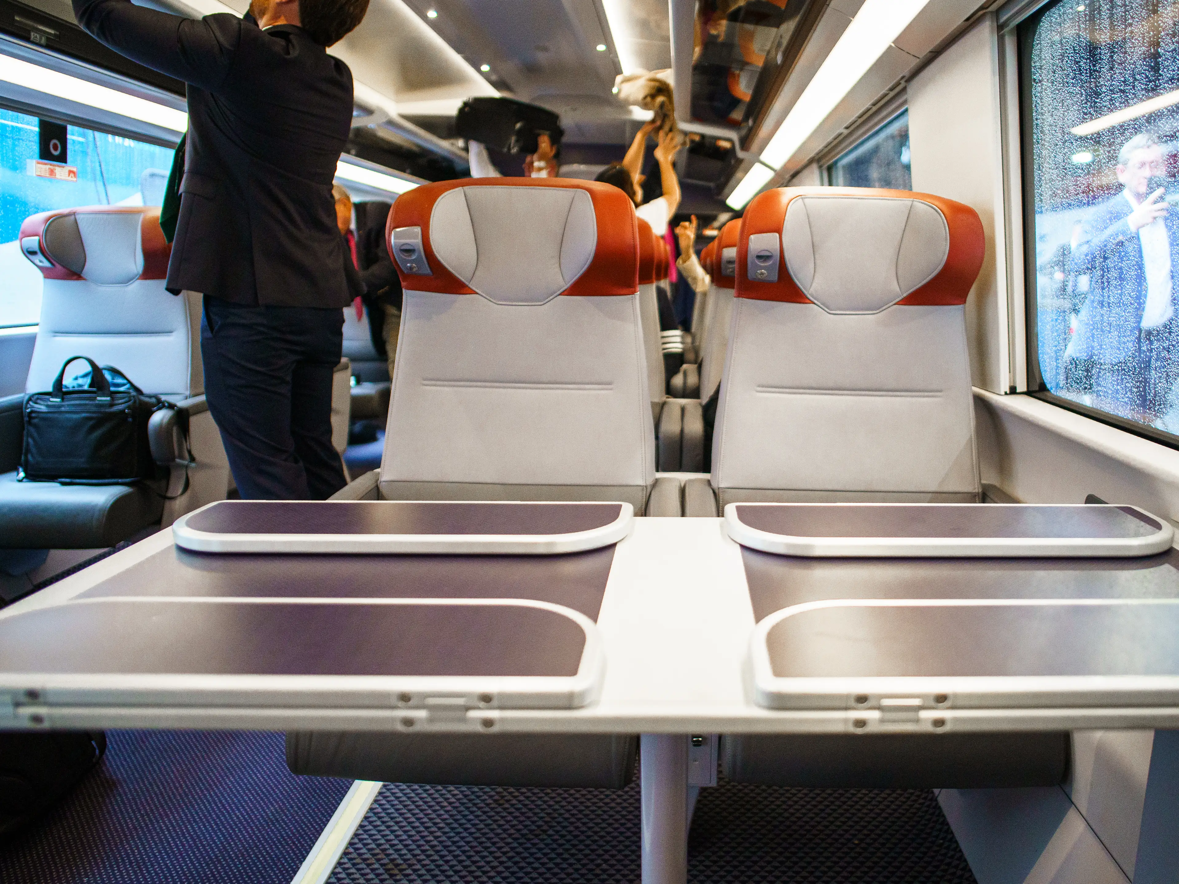 Modern train carriage with paired seats, folding tables, passengers, luggage racks, and rain-speckled windows.