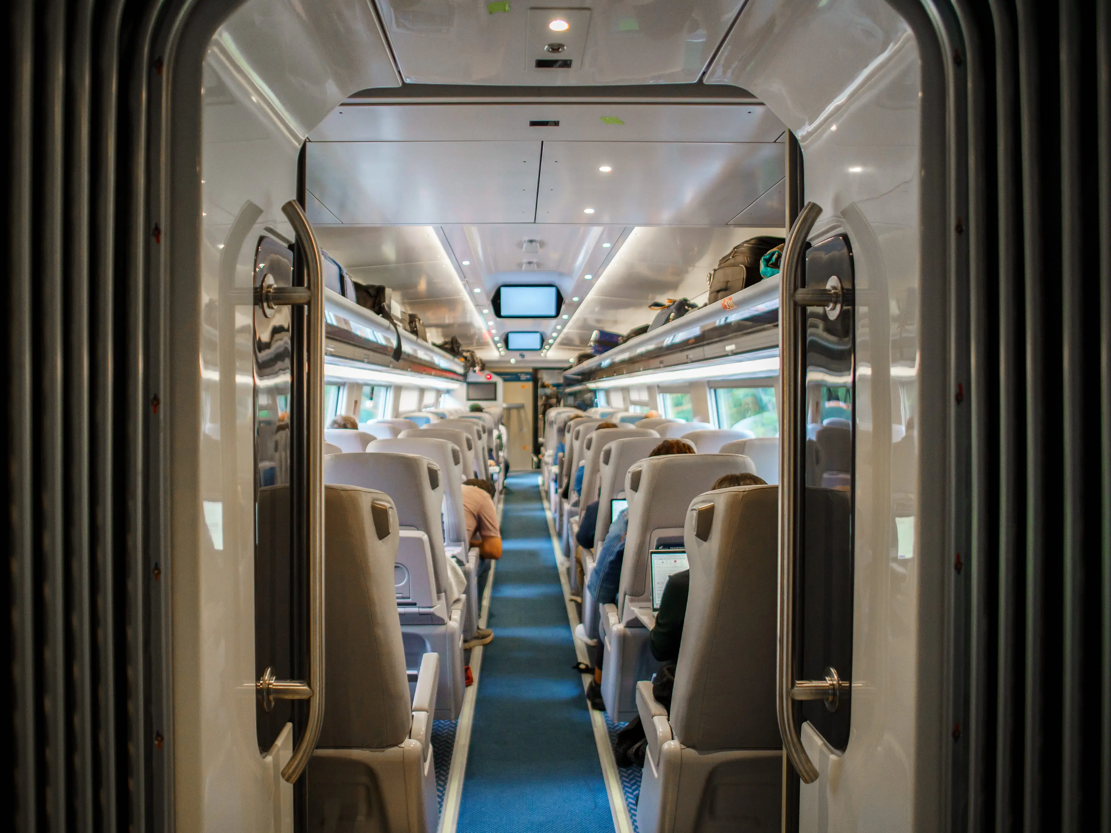 Modern train carriage interior viewed through an open doorway, with rows of seats, passengers, luggage racks, and a blue aisle.