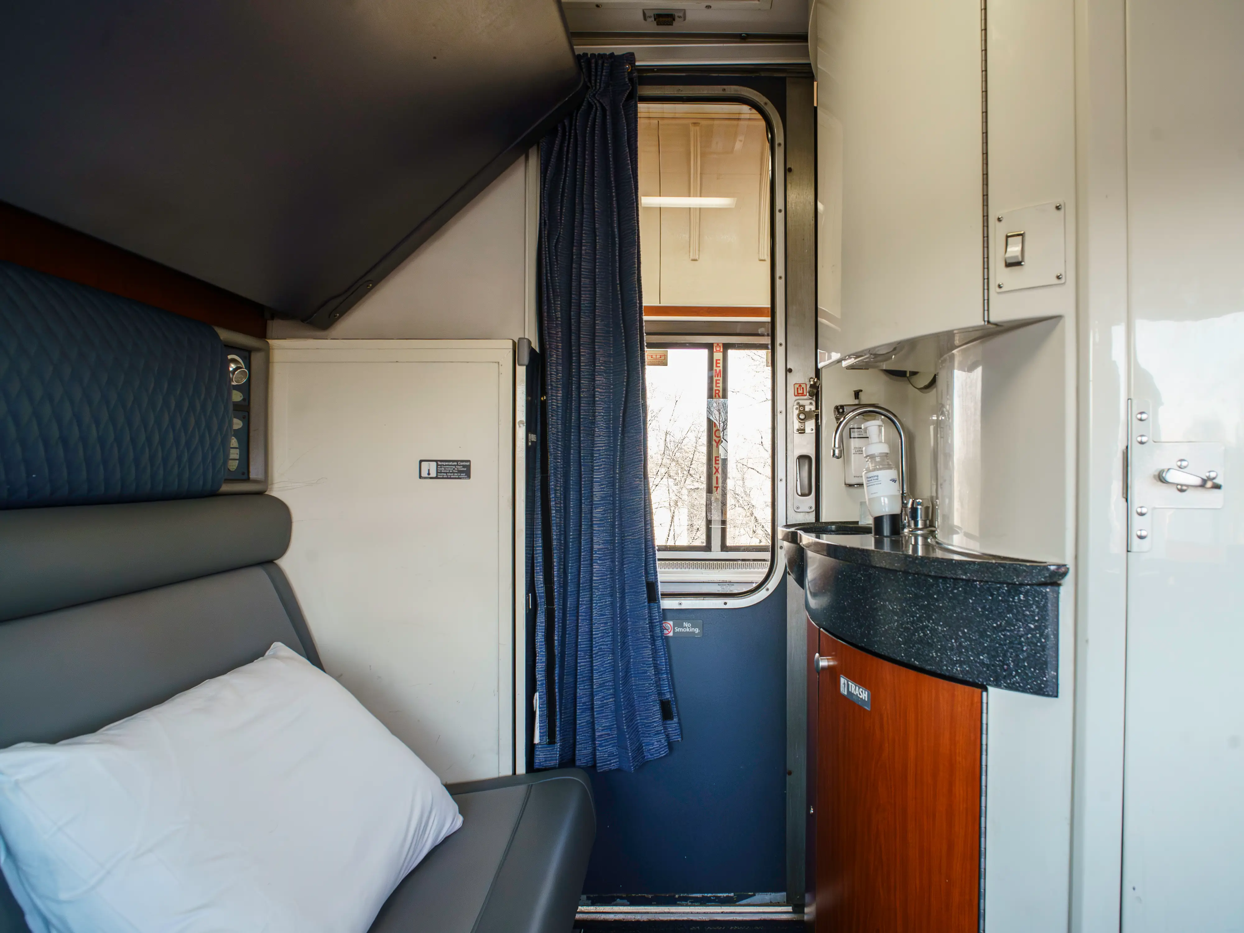 Compact train sleeper compartment with a bed, pillow, blue curtain, windowed door, and small sink area.