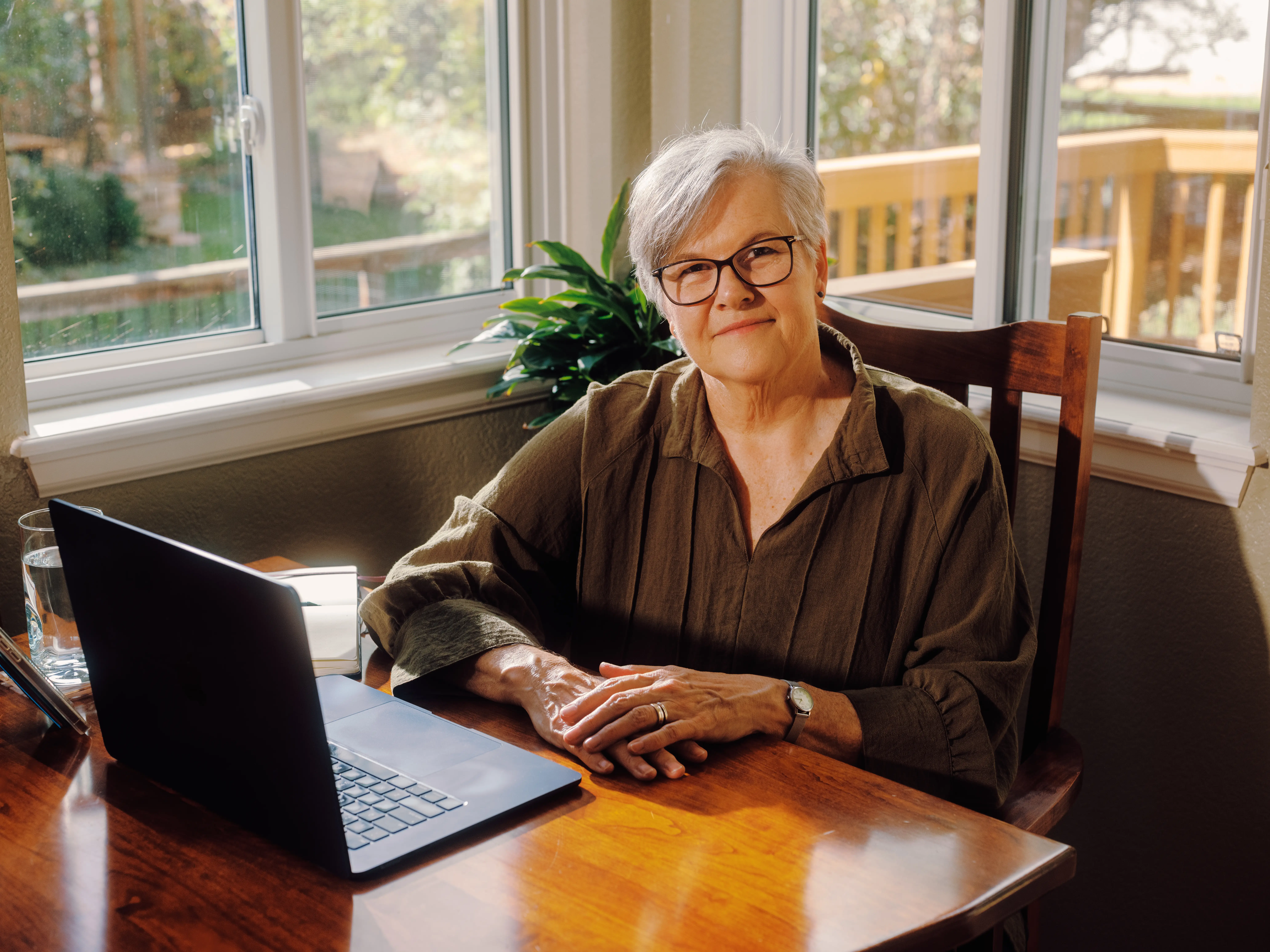 Becky sits at a wooden table beside an open laptop in a sunlit room.