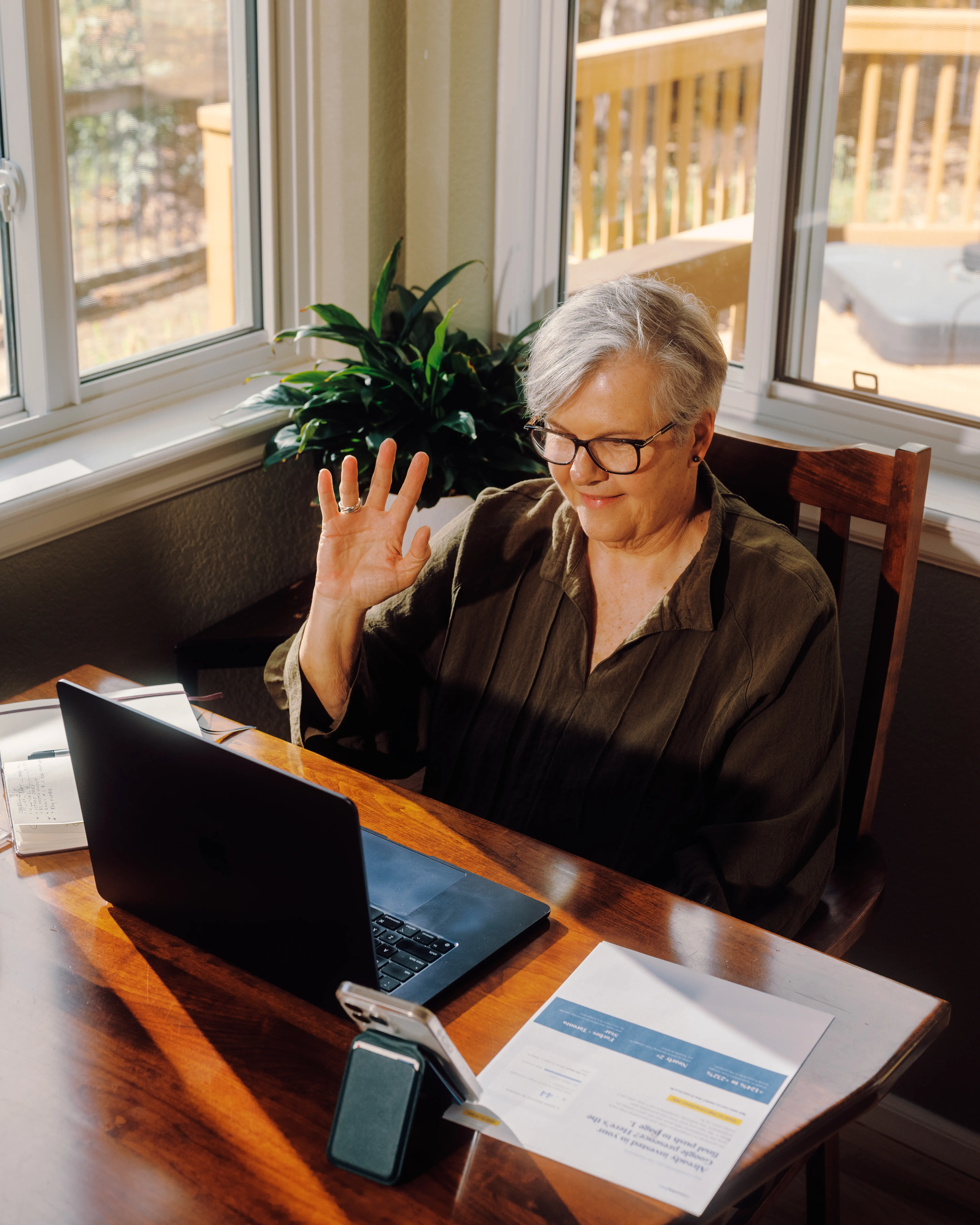 Becky waves while seated at a wooden table using a laptop in a sunny room.