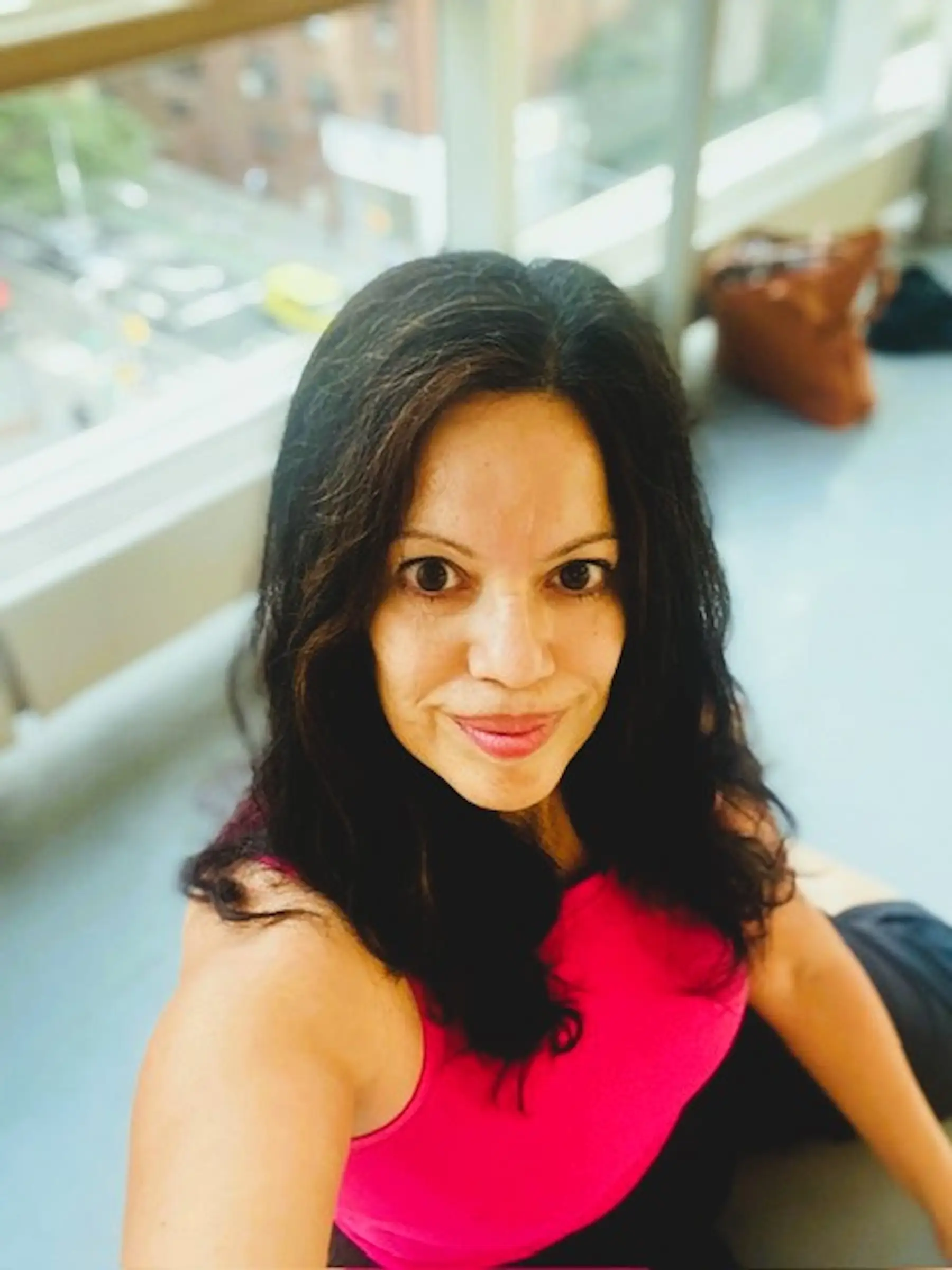 Person in a bright pink sleeveless top takes a selfie beside large windows overlooking a city street.