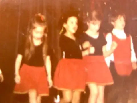 Four performers in coordinated red and black costumes stand on a dim stage beneath a large reflective backdrop.