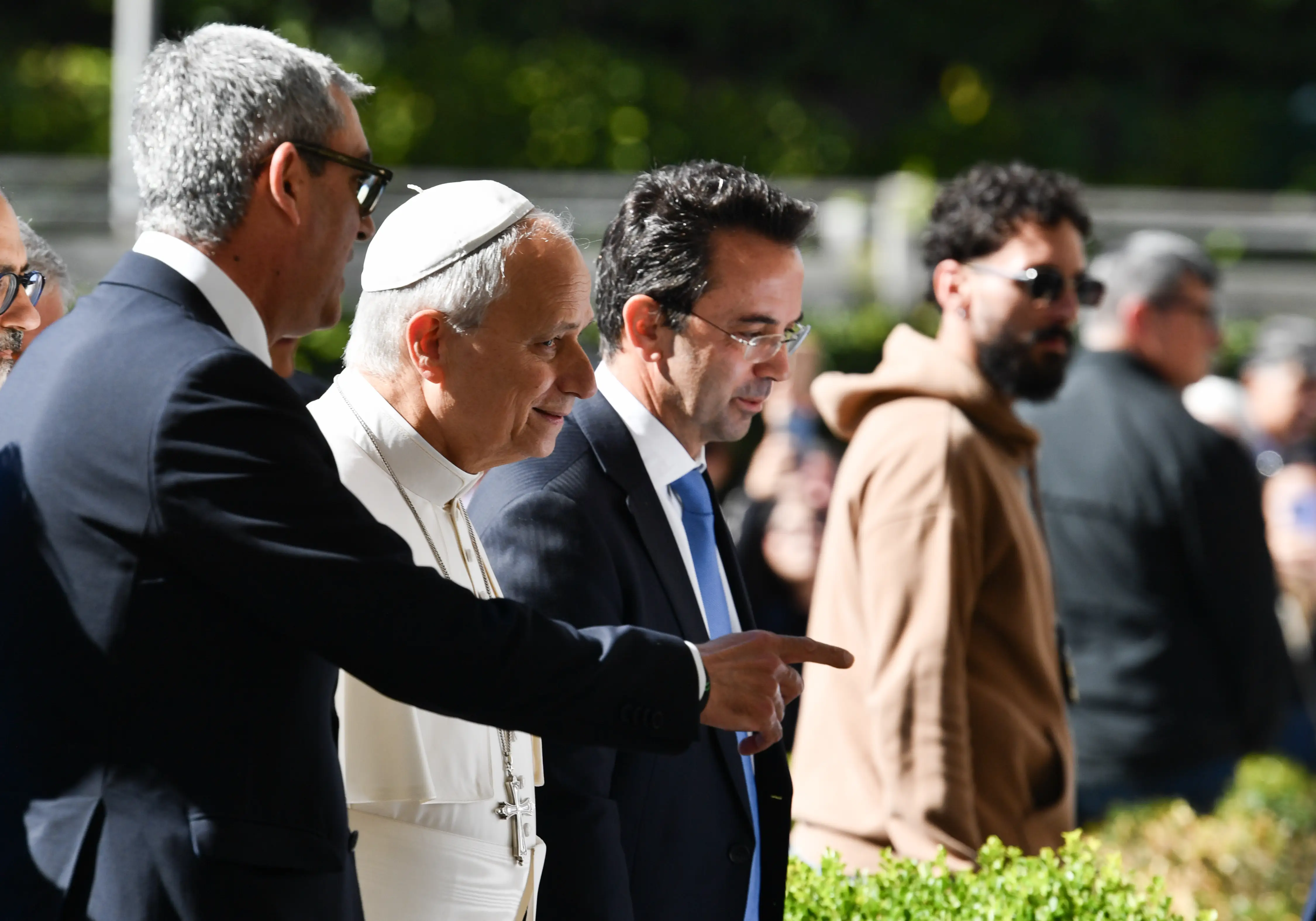 Pope Leo XIV walks outdoors with suited aides and security as one person points ahead.