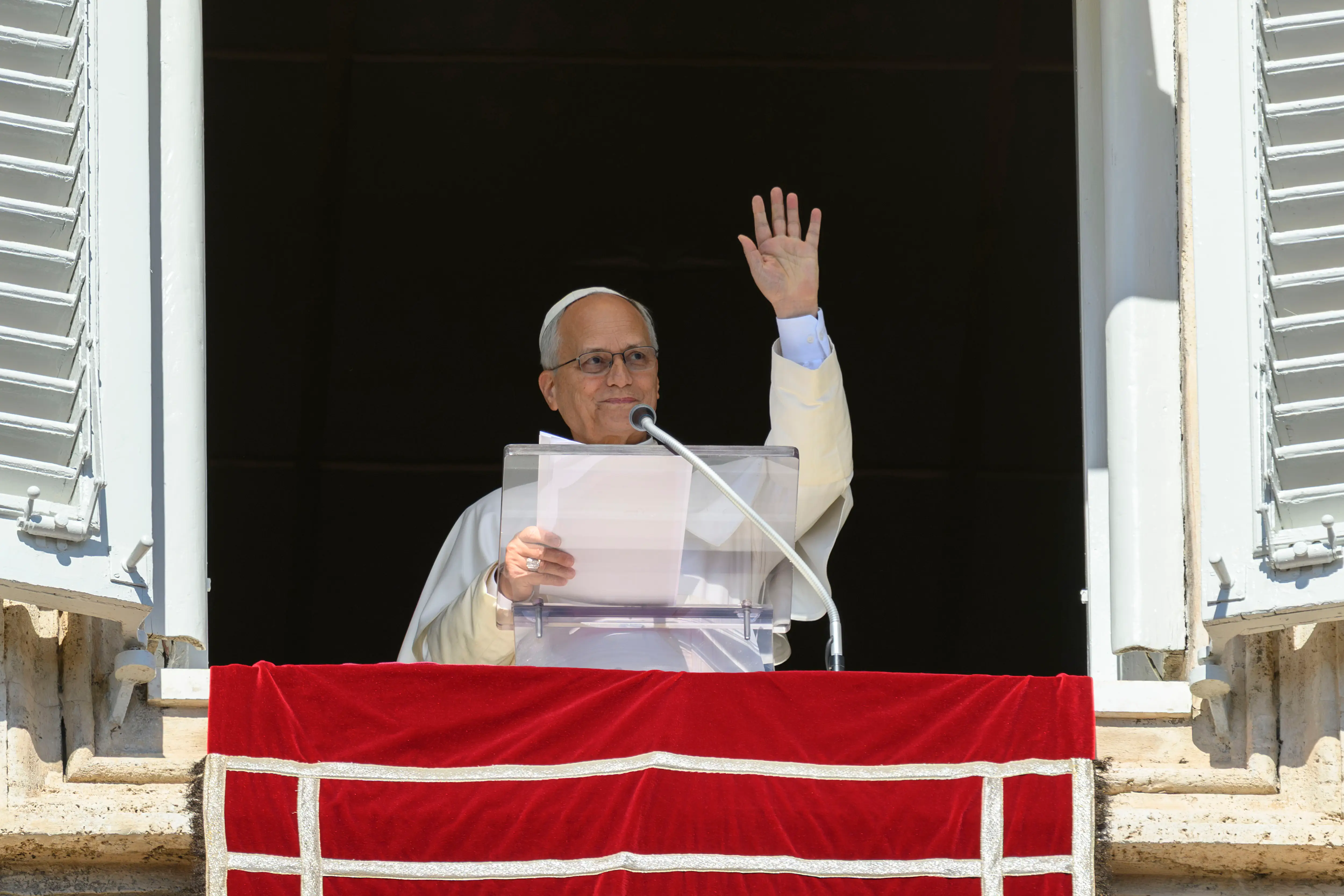 Pope Leo XIV waves from a Vatican window behind a red-draped balcony while holding papers.