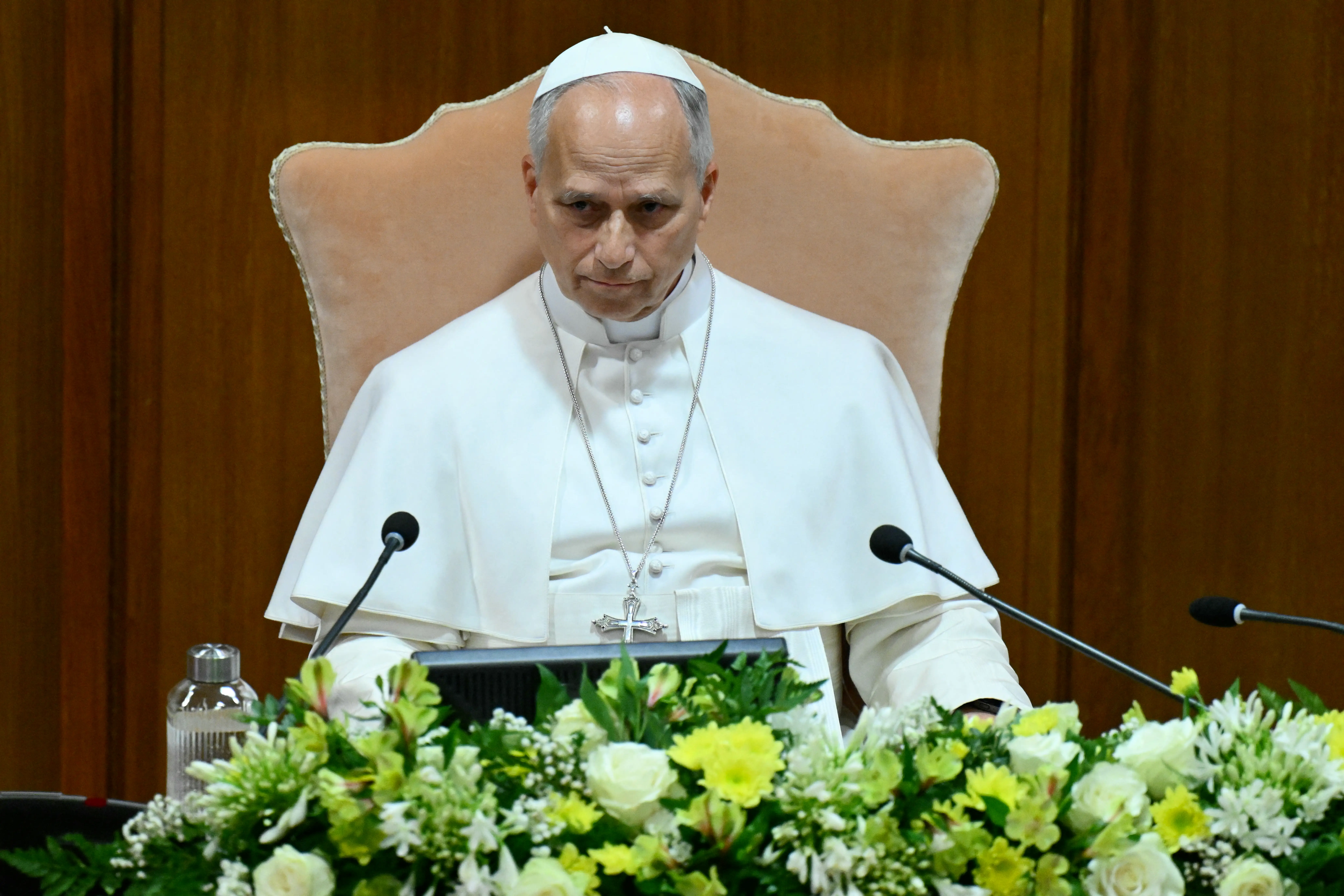 Pope Leo XIV sits at a dais with microphones and flowers against a wood-paneled backdrop.